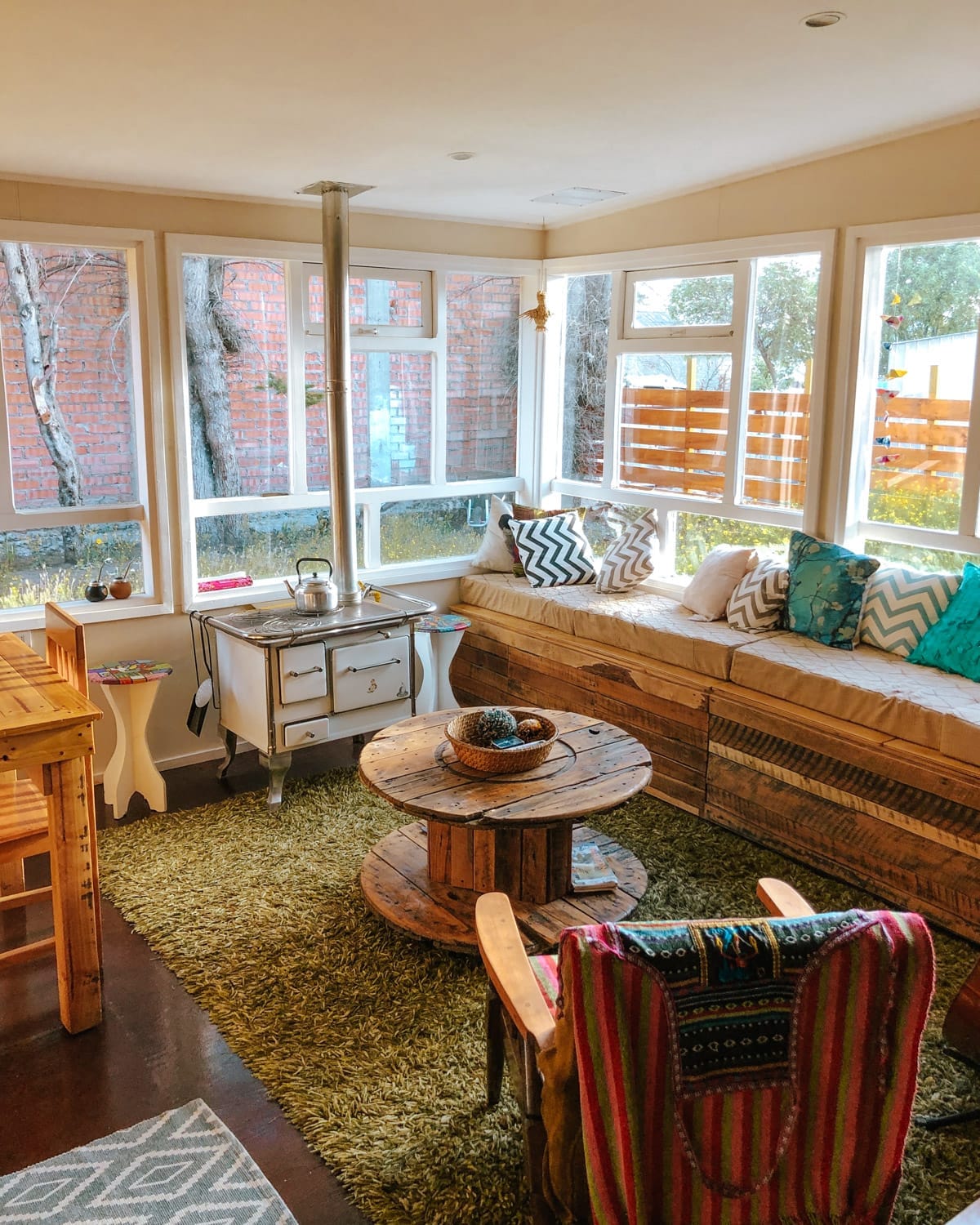 Sunlit common room at Hostal Treehouse with wooden furniture, colourful pillows, and large windows.