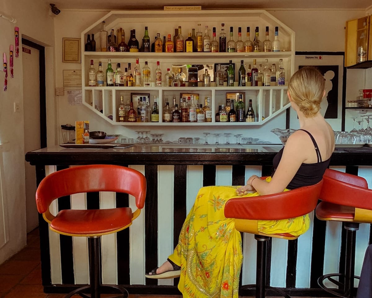 Cec sits at a white and black striped bar counter in St. Lucia with a stocked liquor shelf..