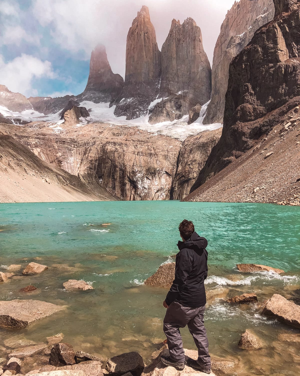 Ari in hiking gear standing by the turquoise lake at Torres del Paine, looking at the three iconic granite peaks.