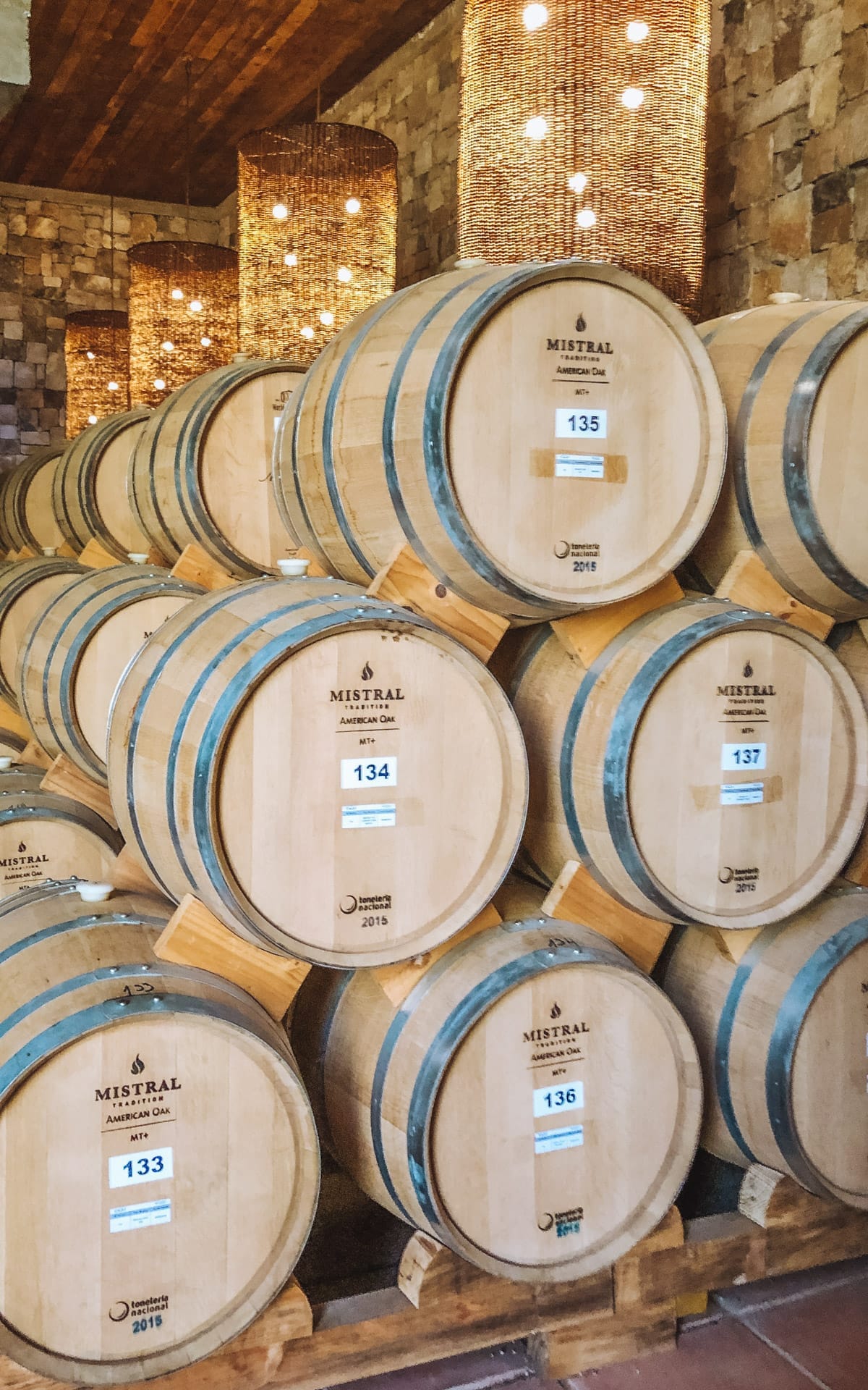 Stacked wooden barrels at the Mistral pisco distillery in Chile, illuminated by hanging beaded chandeliers in a stone-walled room.