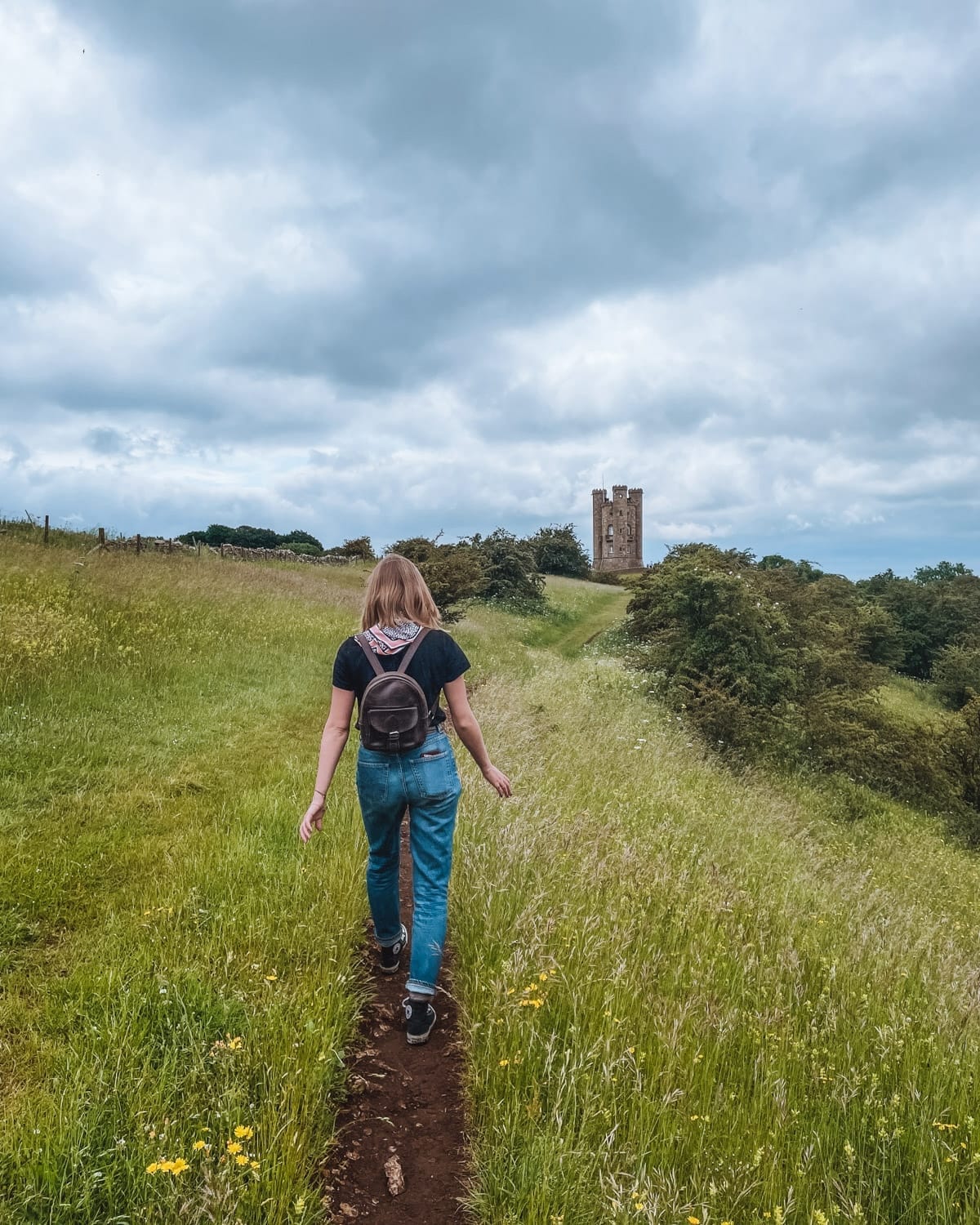 Back view of a Cec's sister walking a narrow trail through green fields toward a distant castle, capturing the charm of the English countryside.