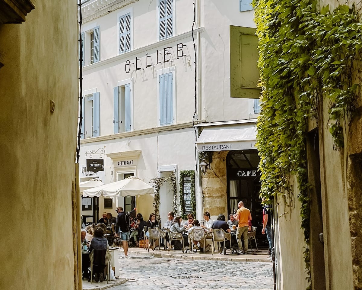 Sunlit alley opening to a café terrace in Provence with people dining beneath umbrellas, evoking classic French café culture.