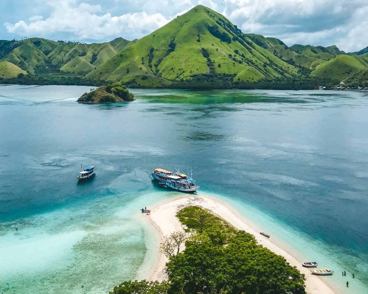 A sandbar with clear turquoise water and boats anchored nearby, set against green rolling hills in Komodo National Park..