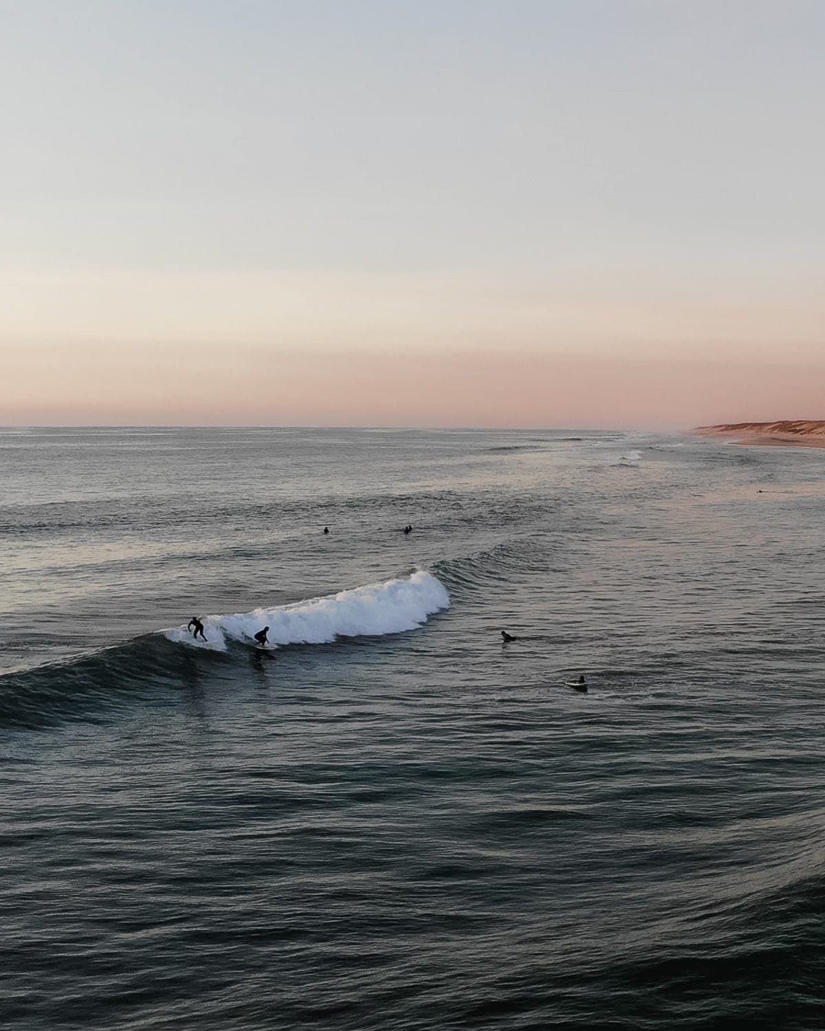 Surfers catching waves at sunset along Cap Ferret’s Atlantic coastline, with pastel hues in the sky reflecting on the water.
