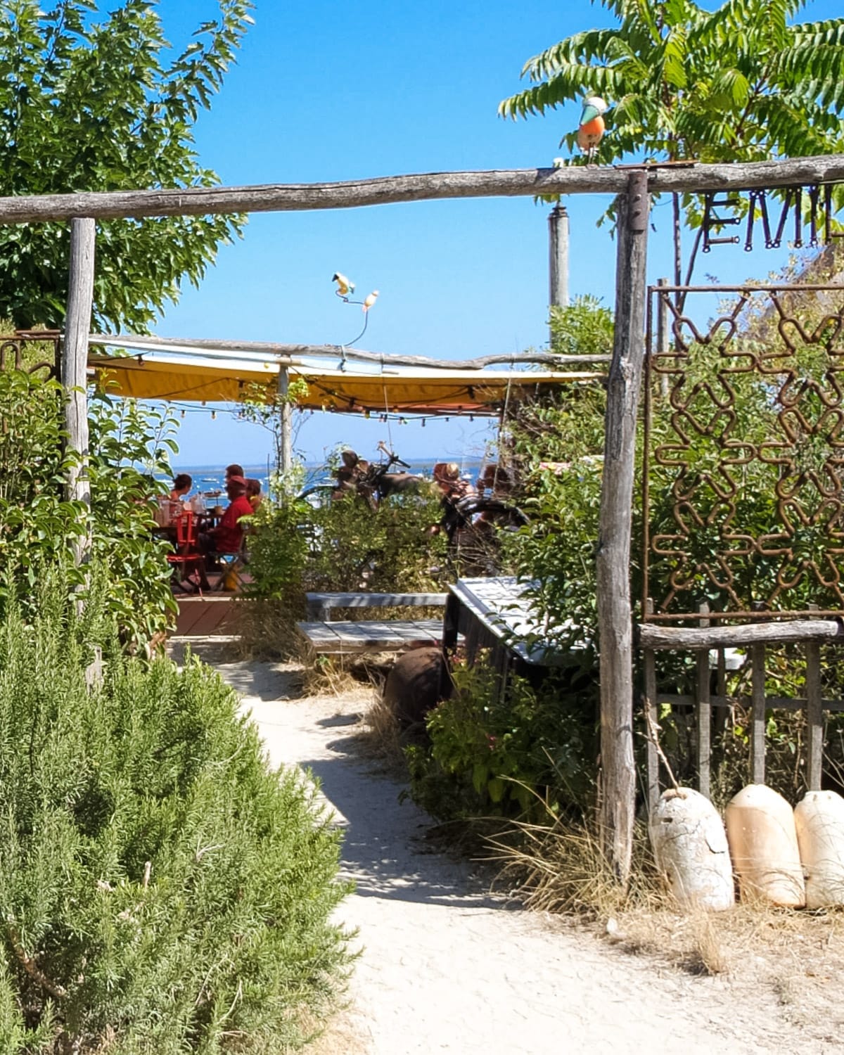 A rustic oyster bar entrance framed by lush greenery, with people dining under a yellow canopy.