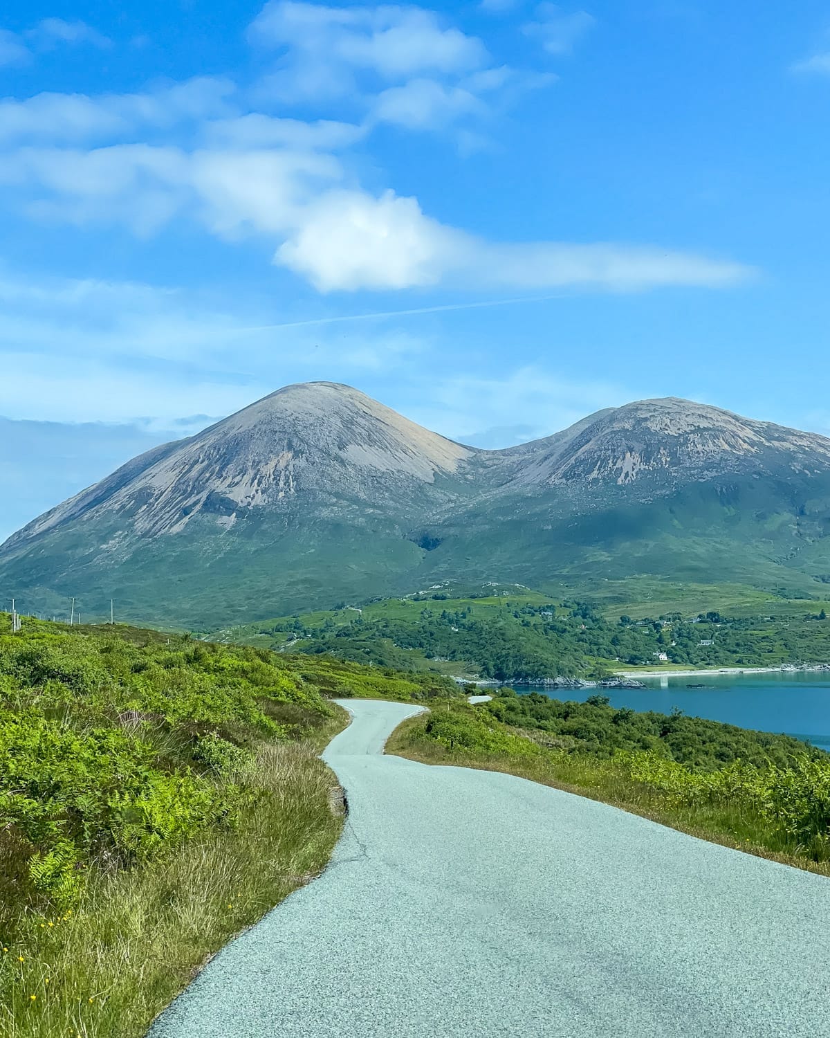 A quiet, winding road leads toward two rounded mountains, with lush green hills and a glimpse of the sea on a sunny day on the Island of Skye.