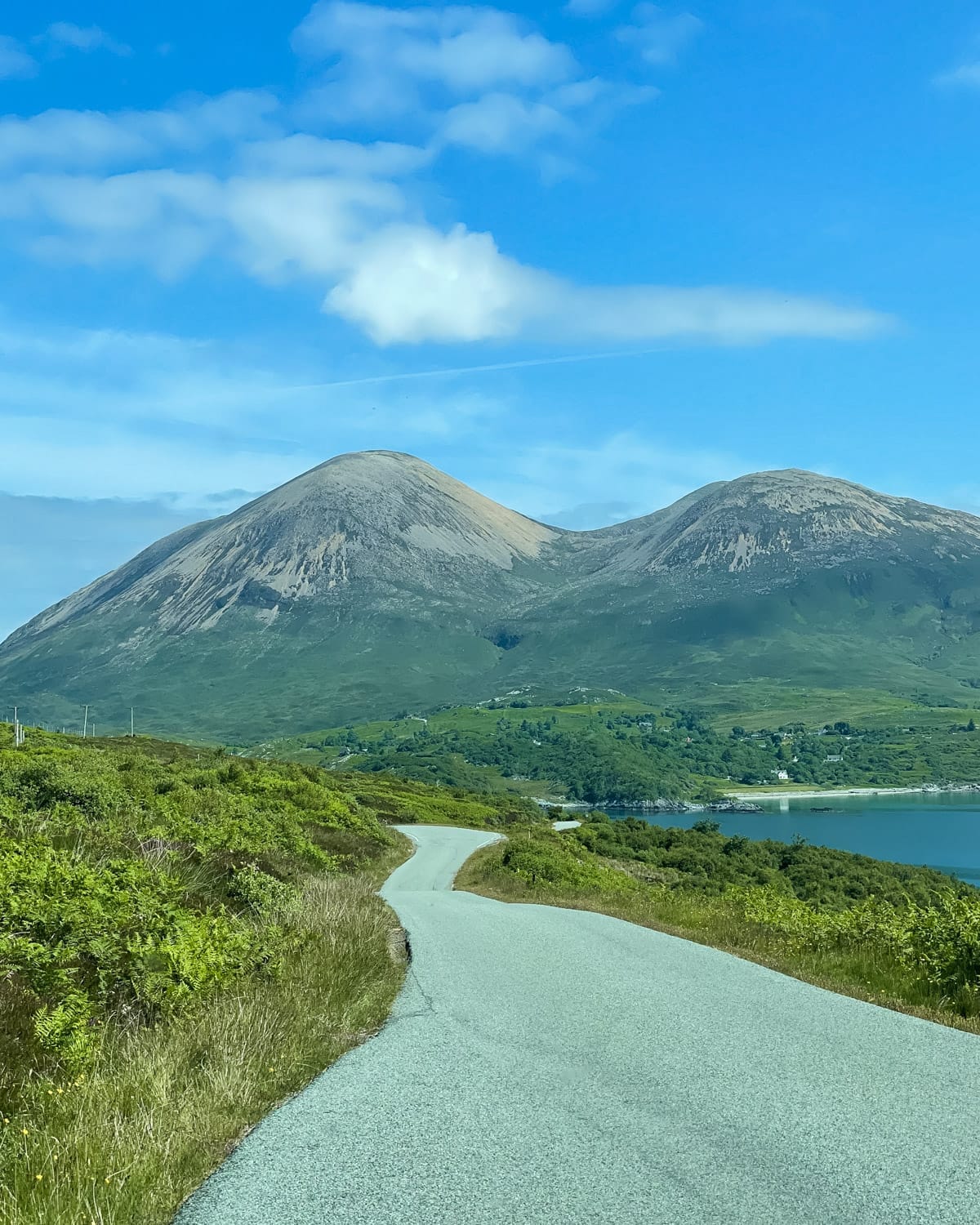 Winding road leads through lush green hills toward the Cuillin Mountains on the Isle of Skye, under a bright blue sky.