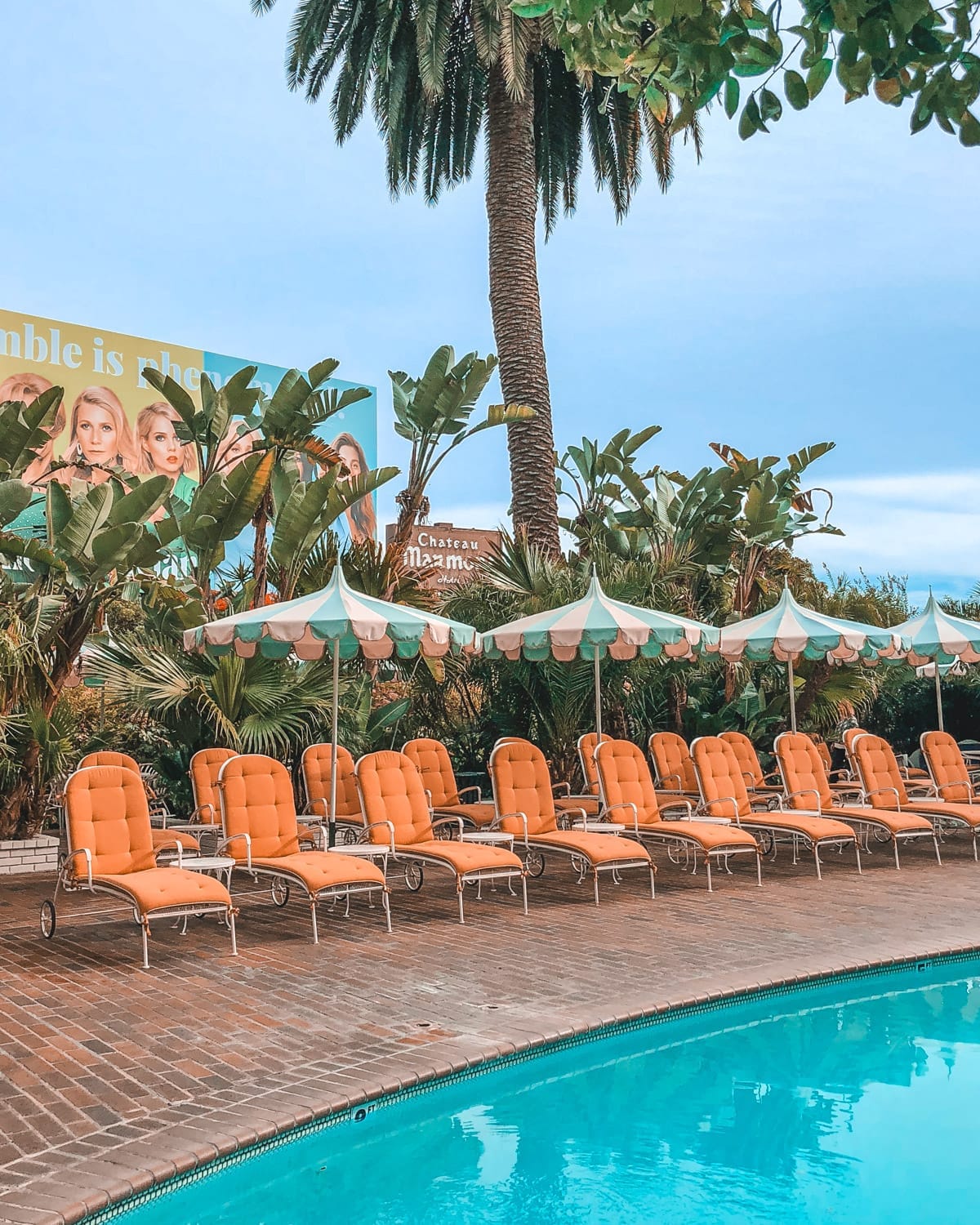 Retro poolside loungers and striped umbrellas at Chateau Marmont in Los Angeles, framed by palm trees and a billboard.
