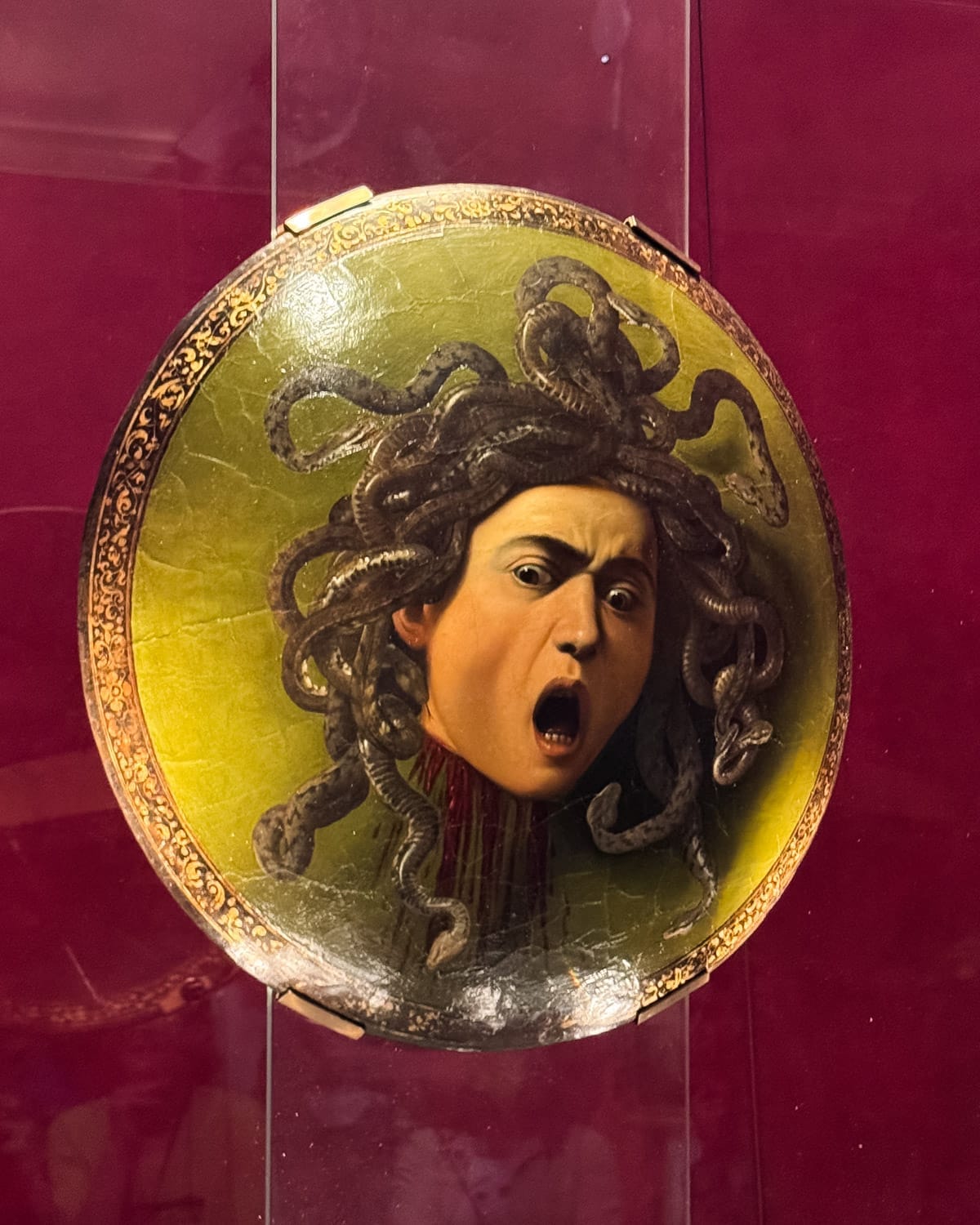 Caravaggio’s ‘Medusa’ shield painting on display at the Uffizi Gallery in Florence, with a deep red backdrop.