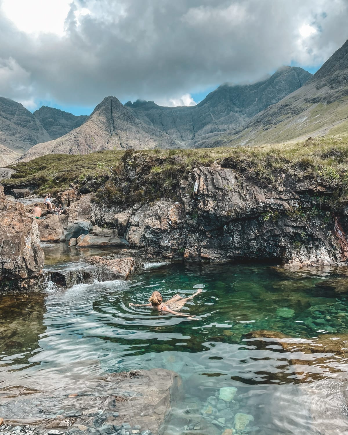 Cec floating in a crystal-clear rock pool surrounded by rugged mountains and moody skies at the Isle of Skye's Fairy Pools. 