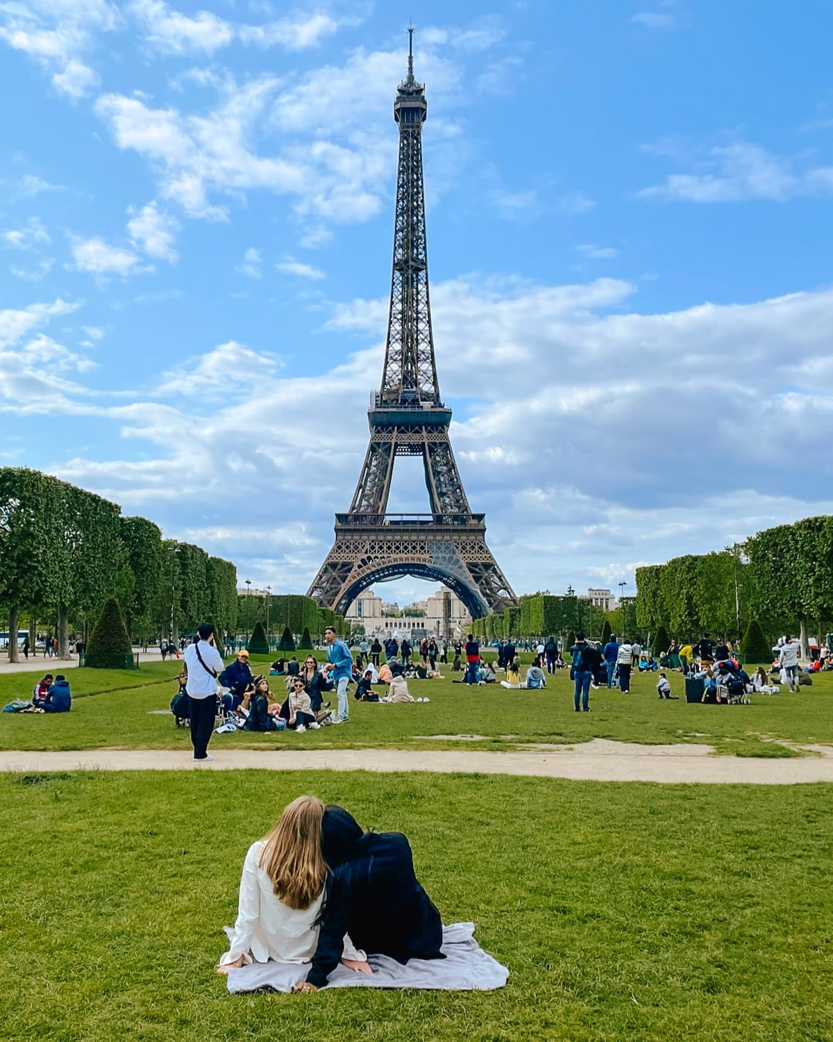Visitors lounge on the lawn beneath the Eiffel Tower in Paris, framed by trees and a blue sky.