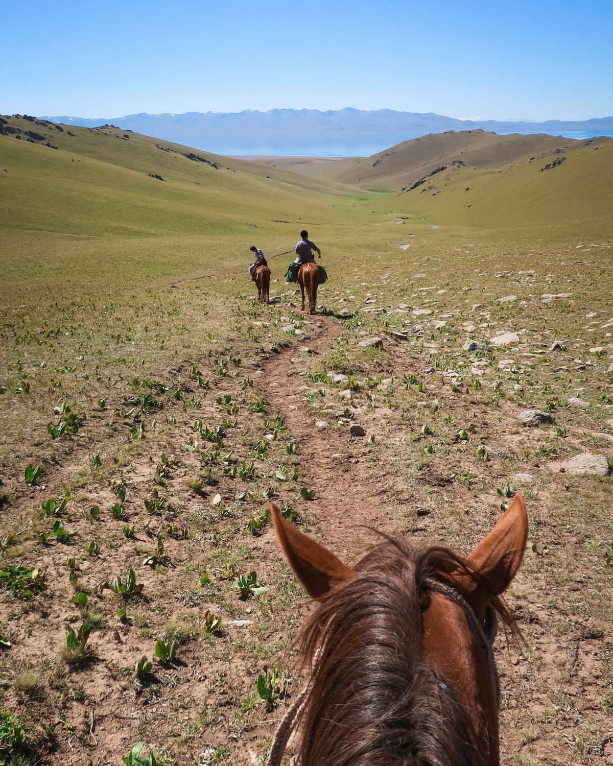 Rider’s view from horseback of two trekkers ahead on a trail through a vast green valley toward the alpine Song Kul Lake in Kyrgyzstan.