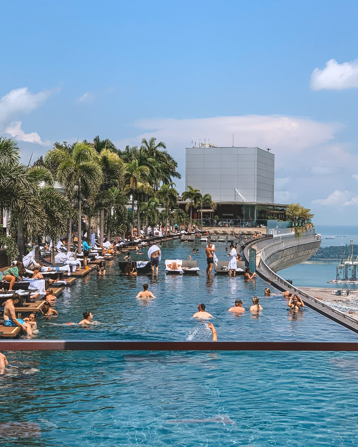 A lively rooftop infinity pool scene at Marina Bay Sands with people swimming and sunbathing under blue skies, overlooking the Singapore harbour.