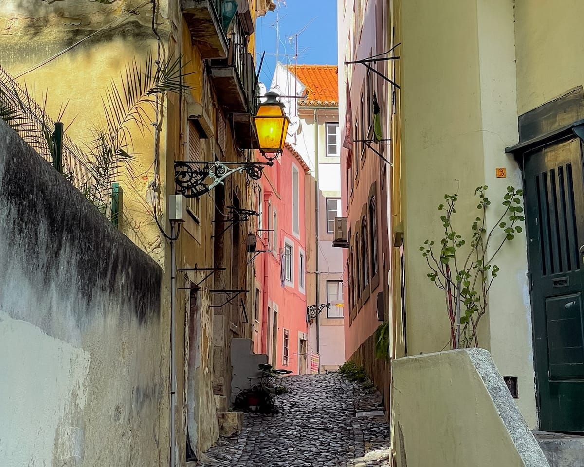 Narrow cobbled alleyway in Alfama, Lisbon, with pastel walls, wrought iron lamps, and warm light streaming in.
