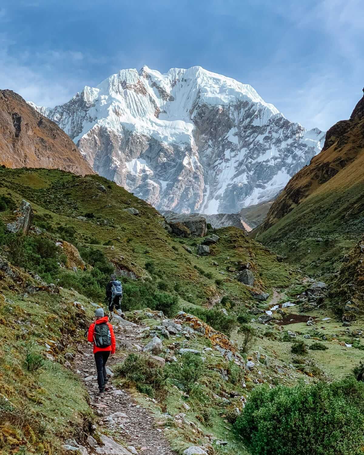 Hikers trek through a green valley toward the snow-capped Salkantay Mountain in Peru, surrounded by rugged Andean scenery.