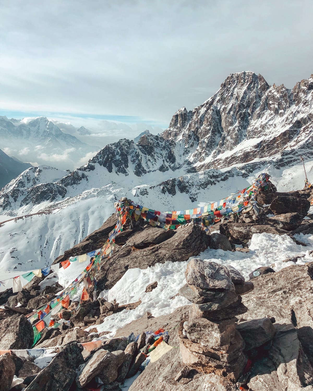Colourful prayer flags flutter on snow-covered rocks at the summit of Gokyo Ri, with Himalayan peaks in the distance.