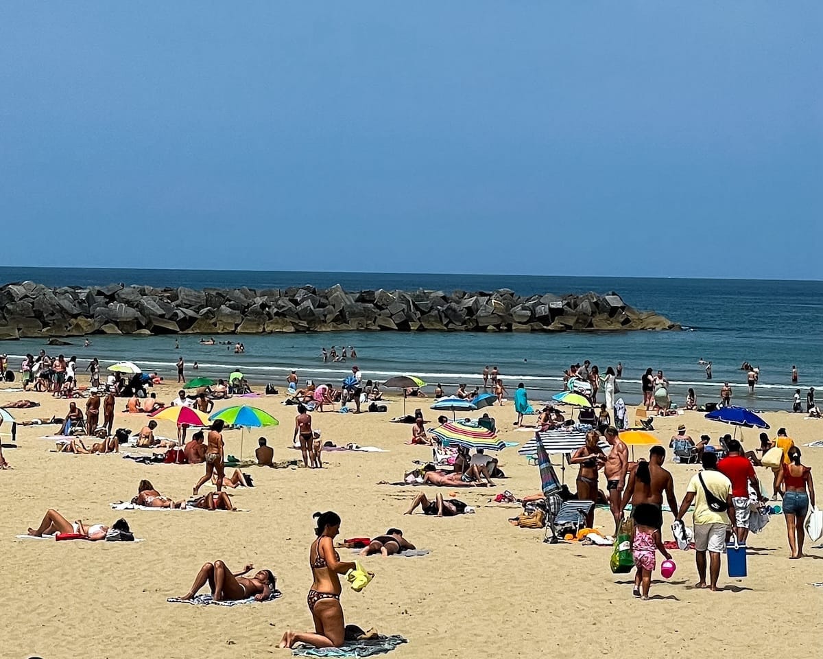 People sunbathing and swimming at a sandy beach in San Sebastian, Spain, with colourful umbrellas and rocky sea break in the distance.