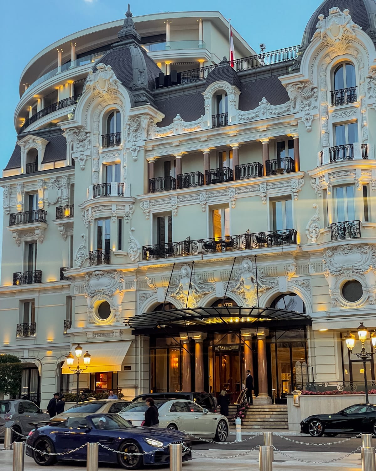 The ornate white façade of Hôtel de Paris Monte-Carlo with luxury cars parked at the entrance during golden hour.