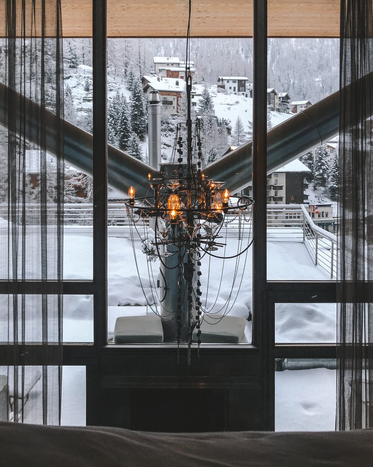 A chandelier hangs in front of a snowy mountain village view in Zermatt, framed by large windows, a bed and thick modern metal beams.