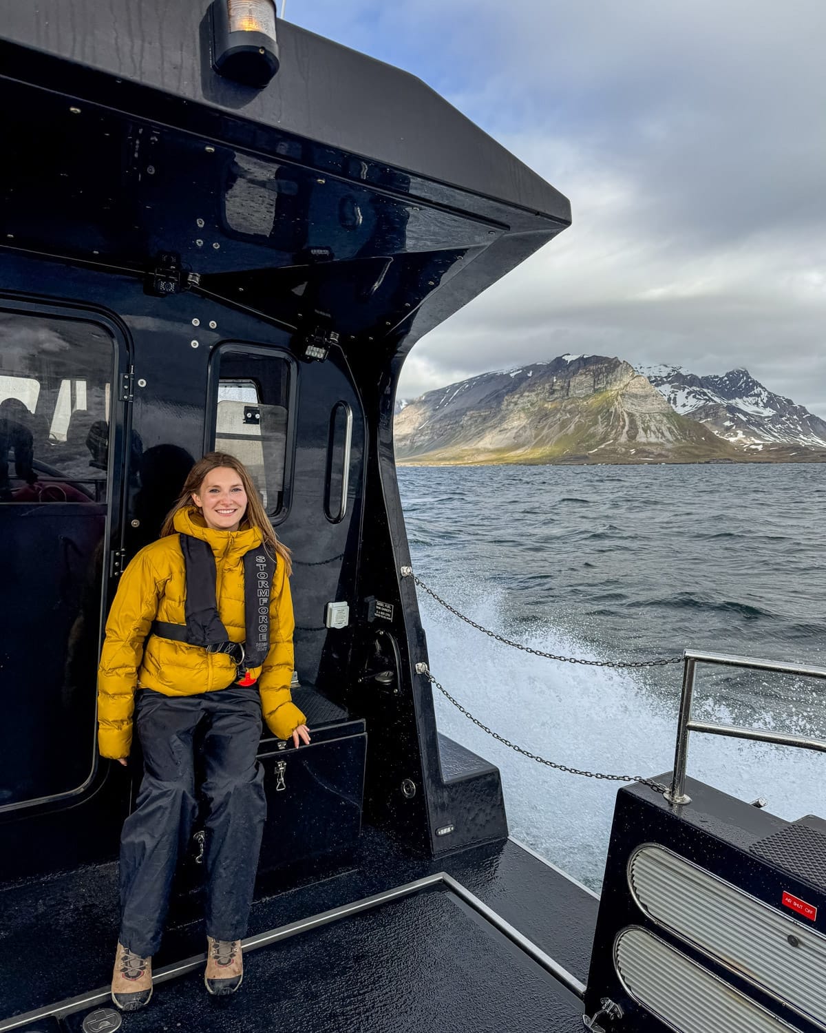 Cec in a yellow jacket rides on a boat near Svalbard with icy mountains rising behind choppy Arctic waters.