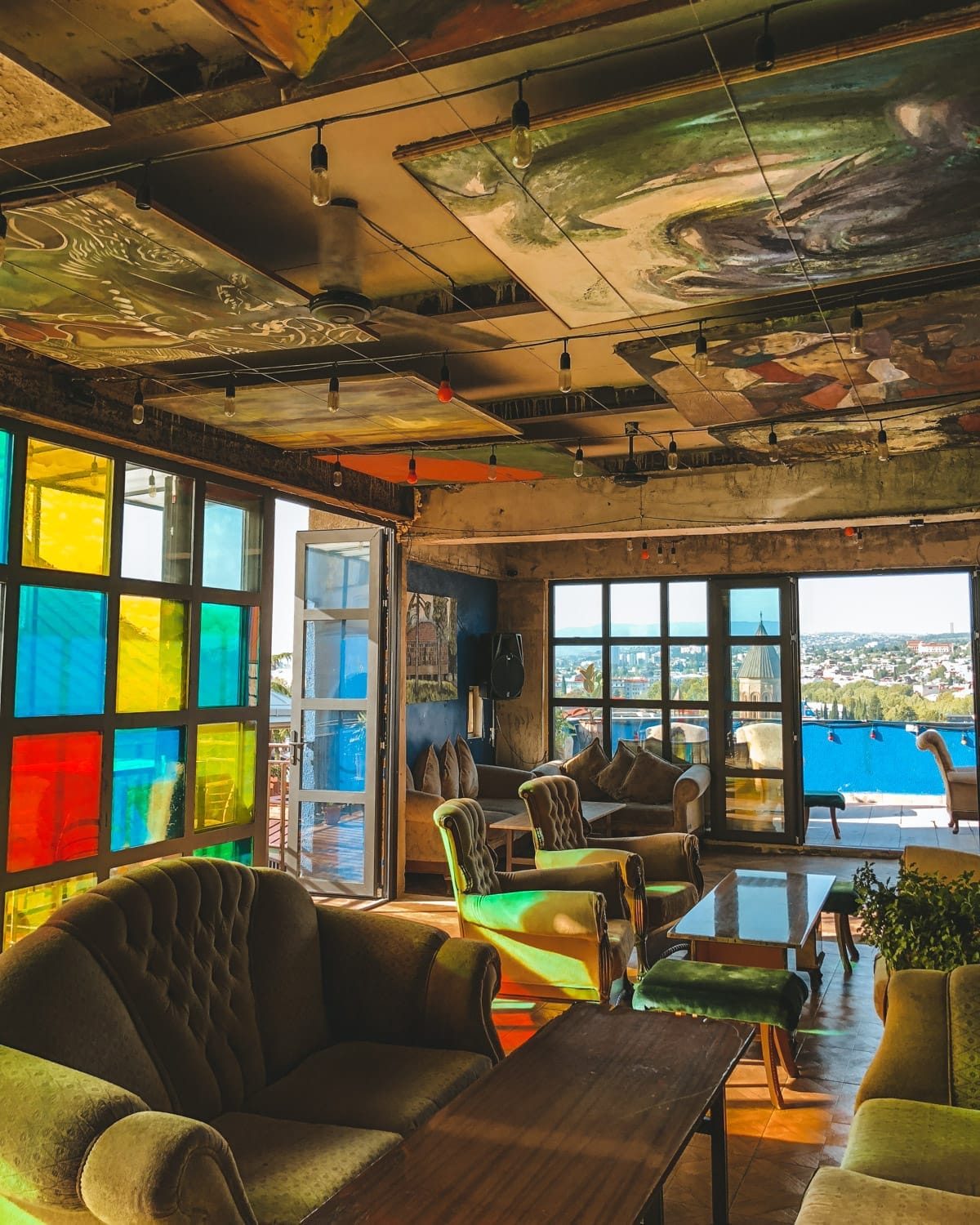 Sunlit art cafe in Tbilisi with ceiling-mounted paintings, vintage furniture, and stained glass windows.