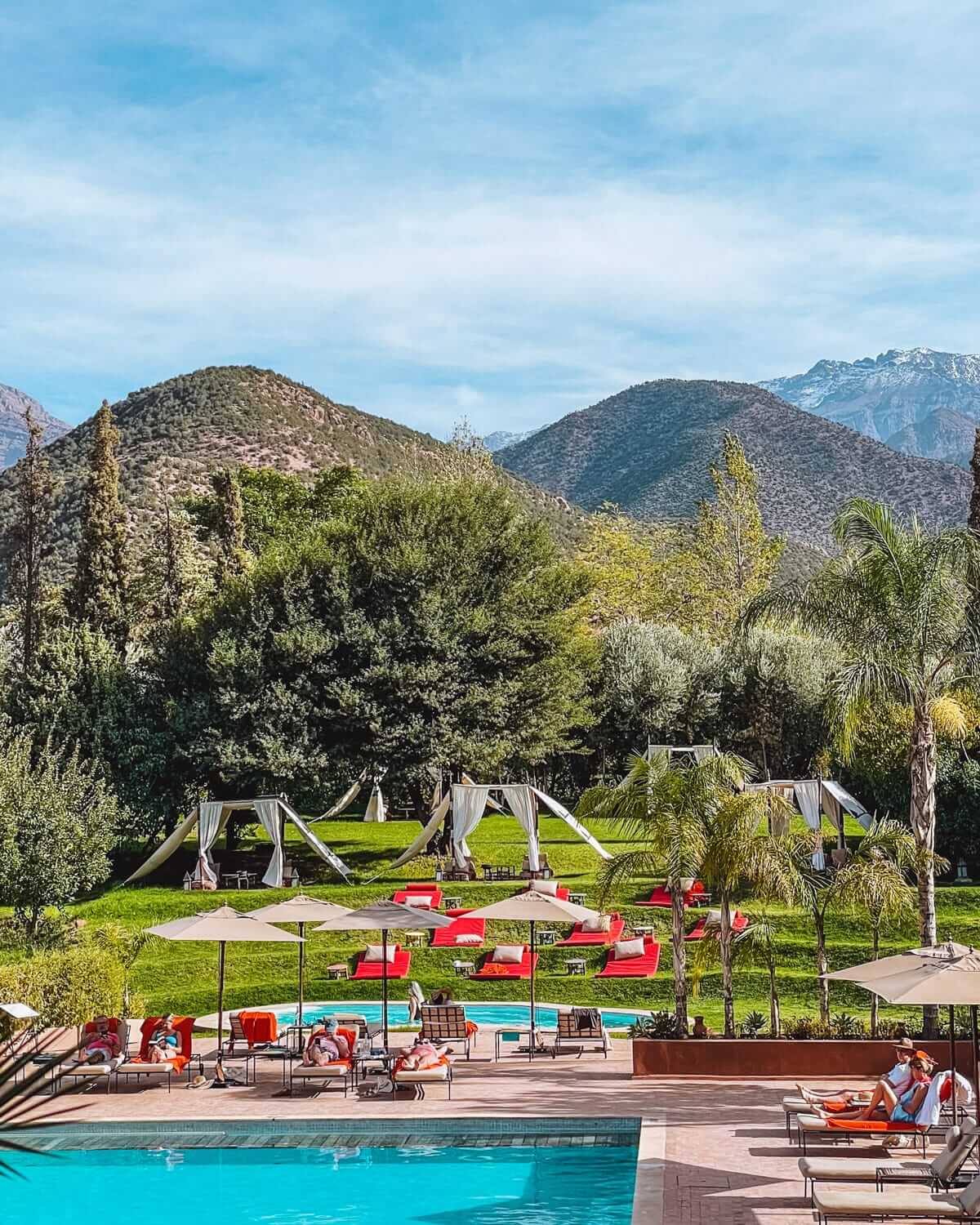 An outdoor pool at a kasbah in the Atlas Mountains, surrounded by red sunbeds, white cabanas, and rounded green mountains.