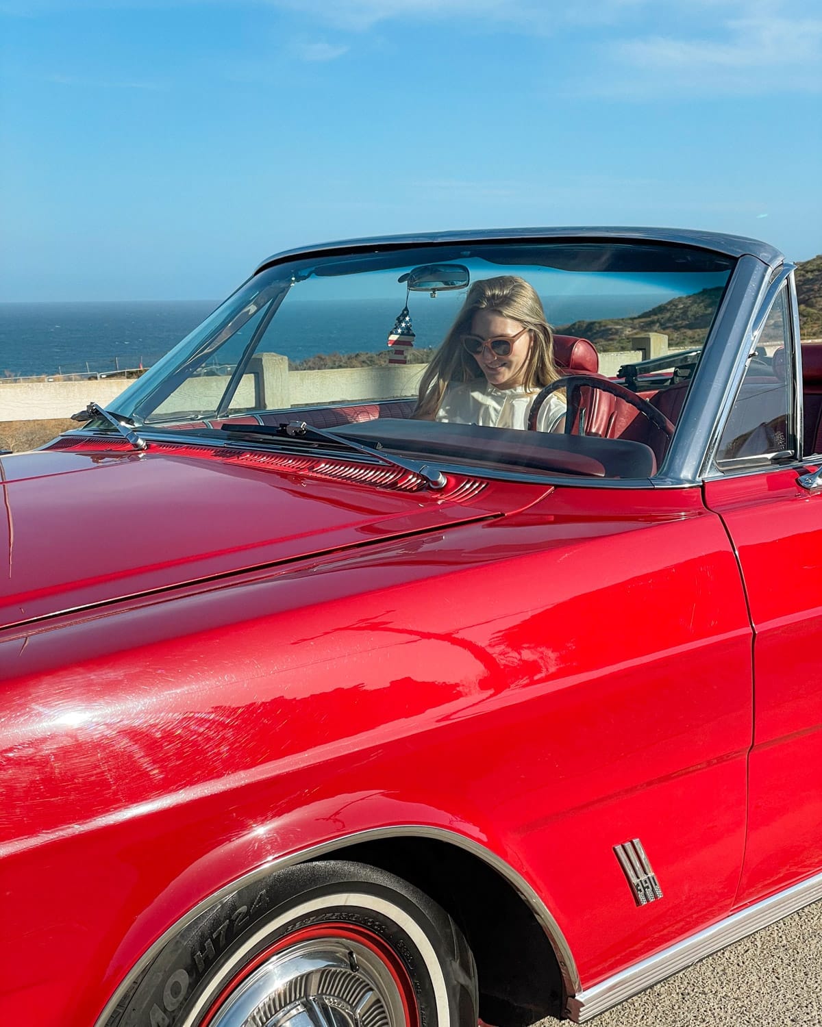 Cec smiles behind in the passenger seat of a red vintage convertible, parked by the Pacific ocean on the California 1 Highway.