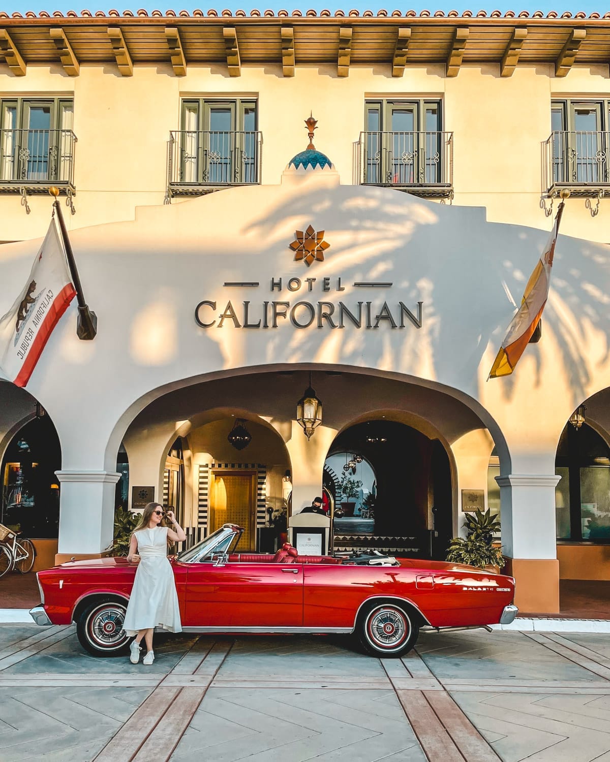 Cec in a white dress stands beside a red vintage convertible parked in front of the Hotel Californian's grand entrance.