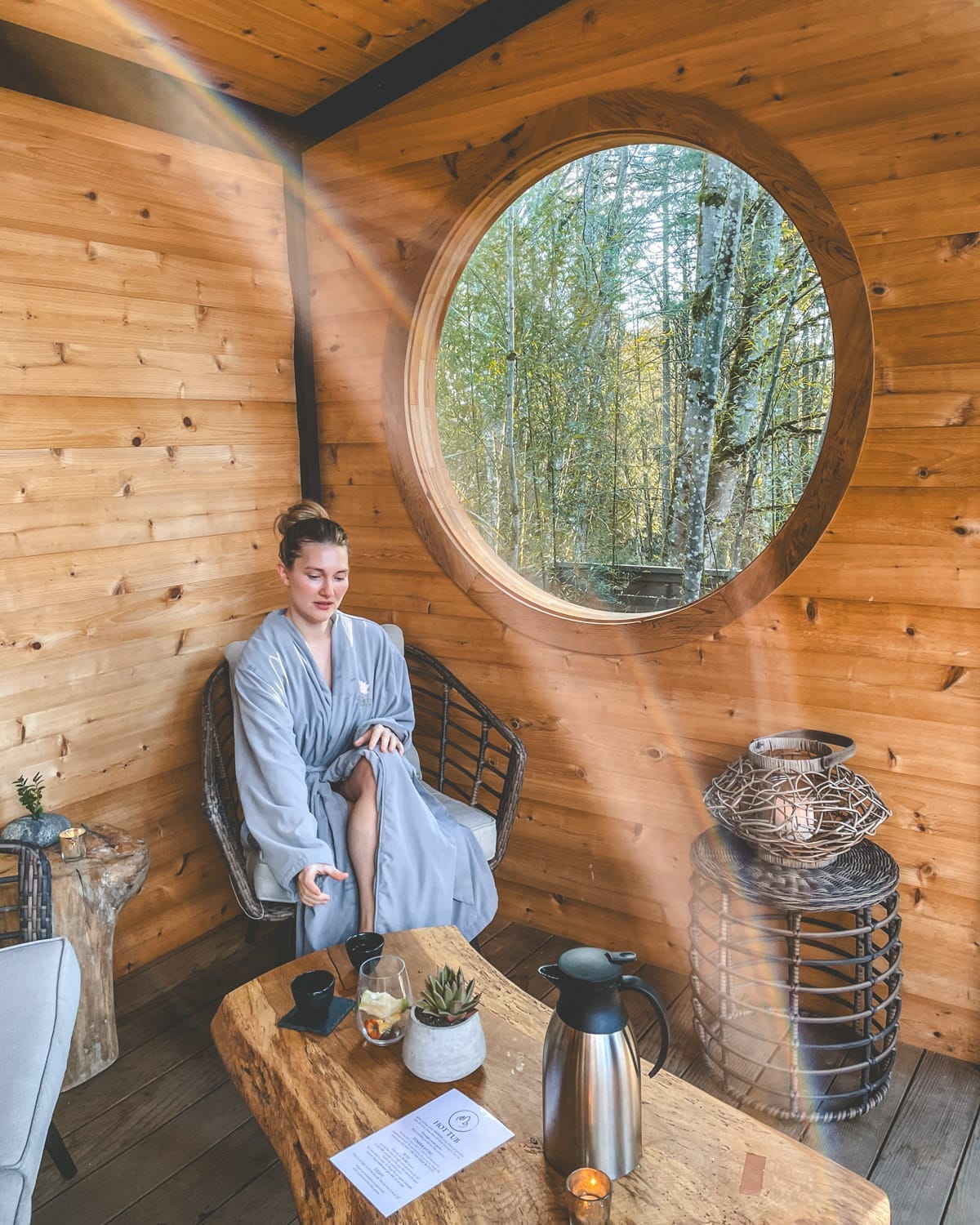 Cec in a robe relaxing in a wooden spa room with a round window, hot tea on the table, and soft sunlight filtering in.