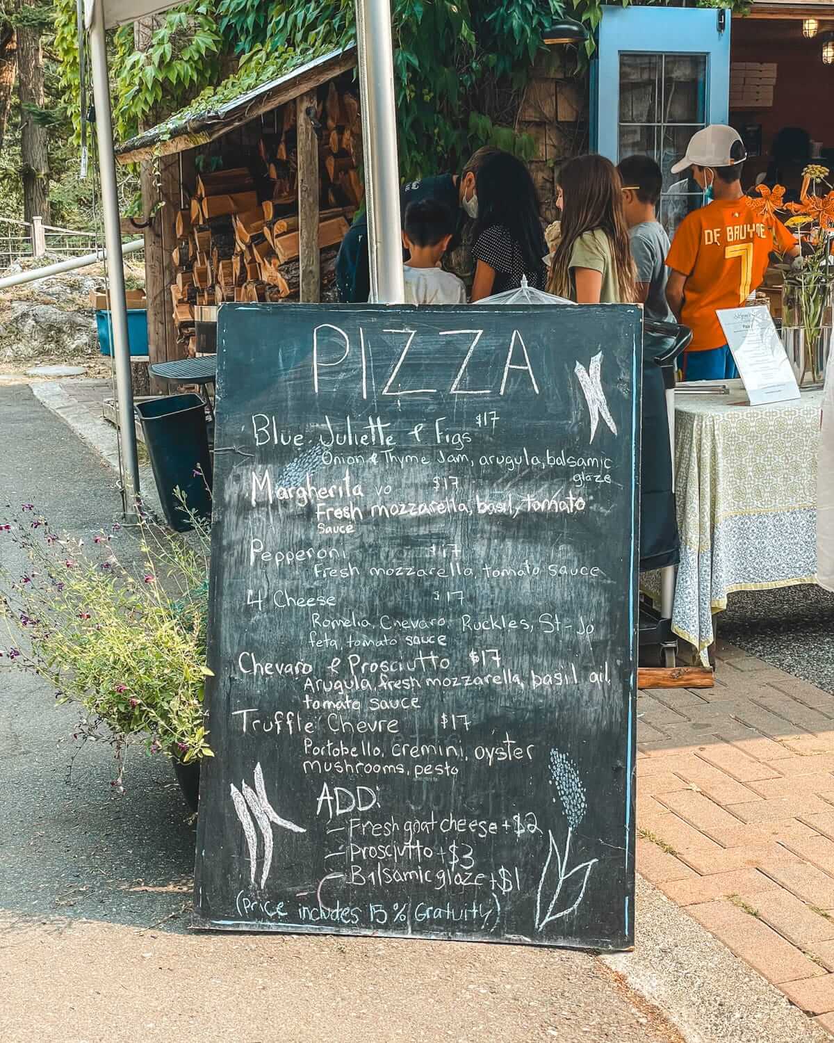 Chalkboard pizza menu at Salt Spring Island Cheese's outdoor pizzeria, listing creative pizza options.