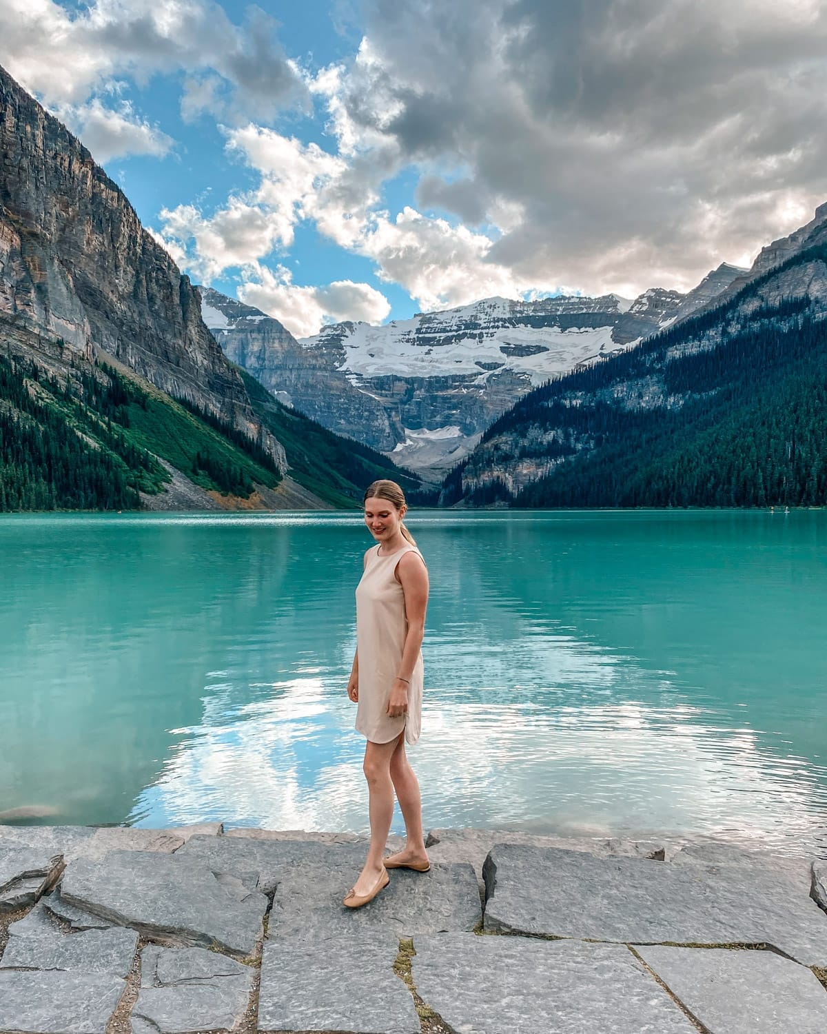 Cec in a peach dress stands on a rocky shore in front of turquoise Lake Louise, surrounded by forest and glacier peaks.