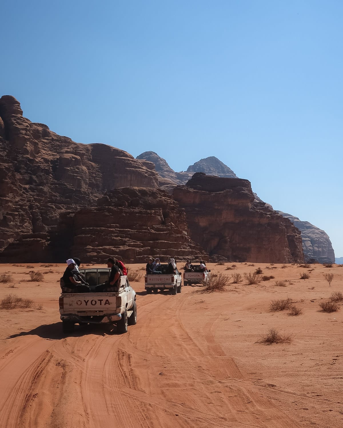 Vintage Toyota trucks with people in the back drive across the orange sand and dramatic rock formations of Wadi Rum under a clear desert sky.