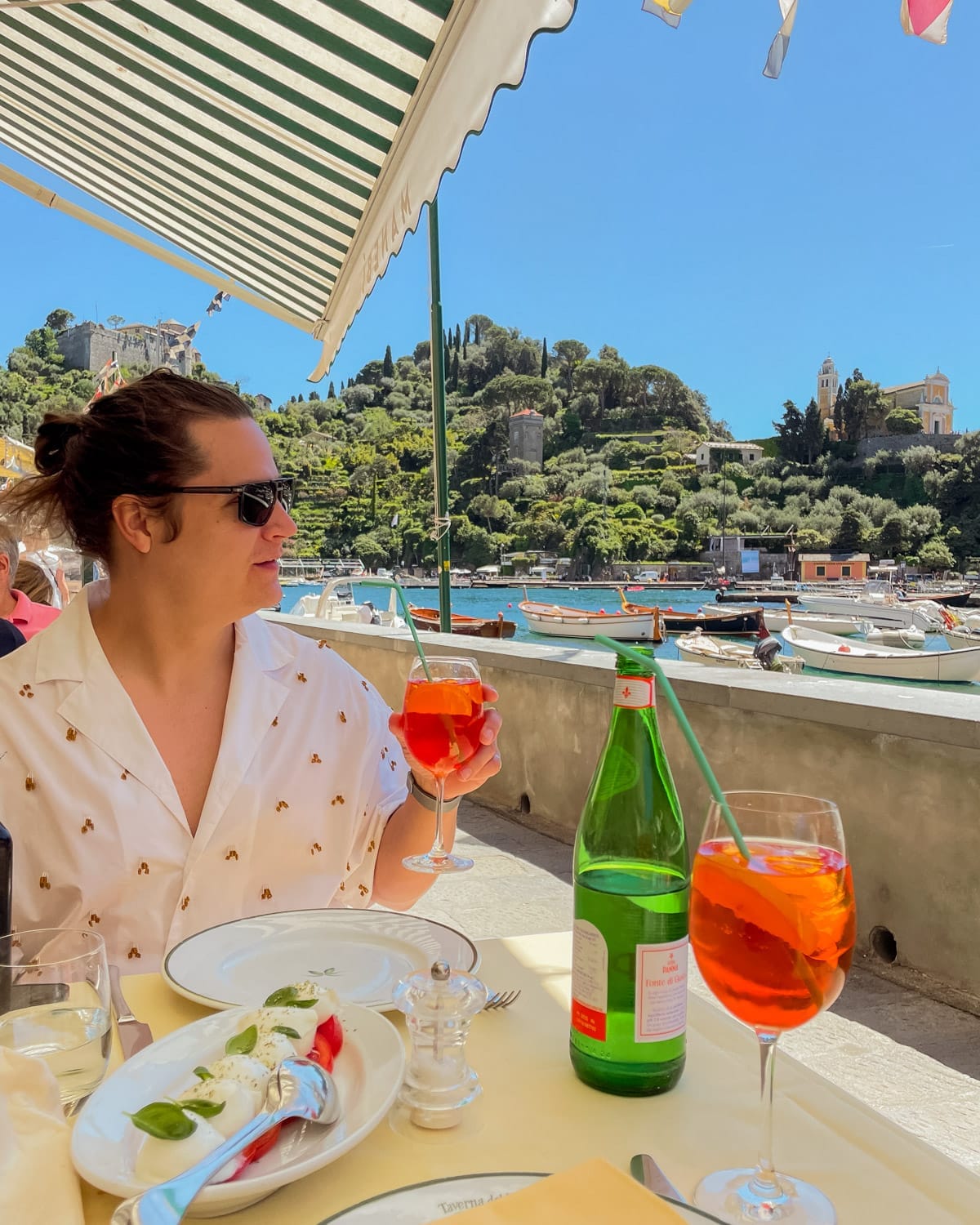 Ari in sunglasses enjoys a bright Aperol spritz and a plate of caprese salad at a harbourside restaurant in Portofino.