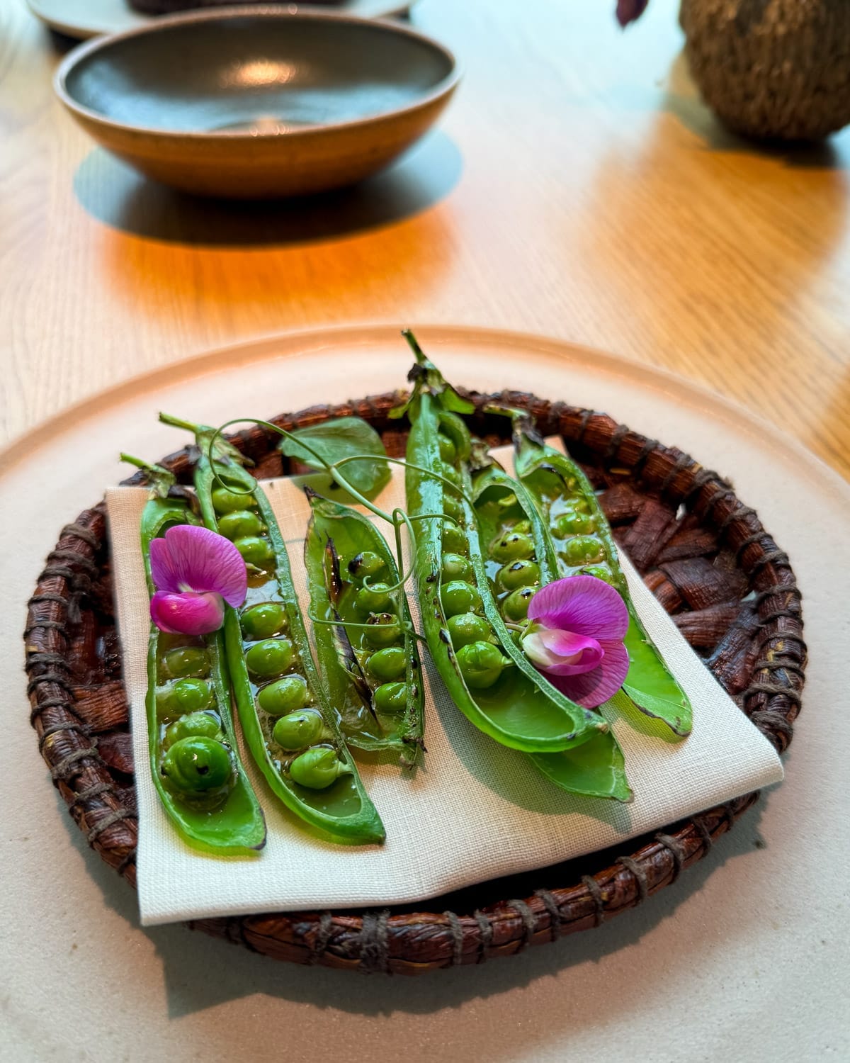 Grilled pea pods garnished with edible flowers, presented on a cloth napkin in a woven dish on a fine dining table at Noma, Copenhagen.
