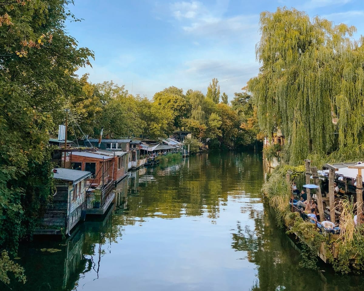 Charming houseboats and waterside cafés line a calm canal surrounded by trees in Berlin.