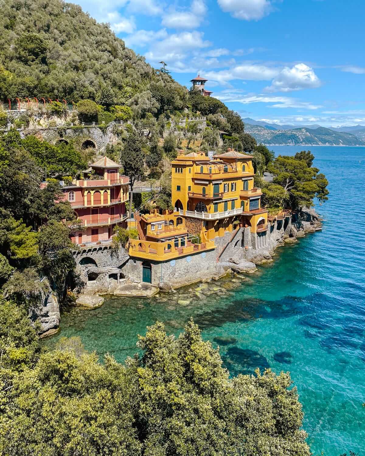Colourful villas perched on rocky cliffs above turquoise waters in Portofino, Italy, surrounded by lush green hills.
