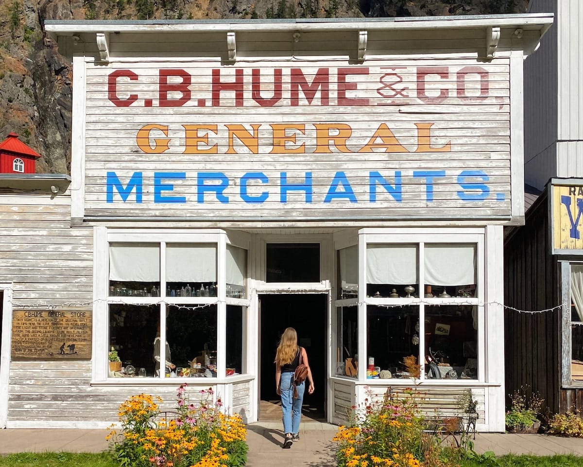 Traveler enters a general merchants shop at Three Valley Gap's historic ghost town, framed by rustic white wood and blooming flowers.