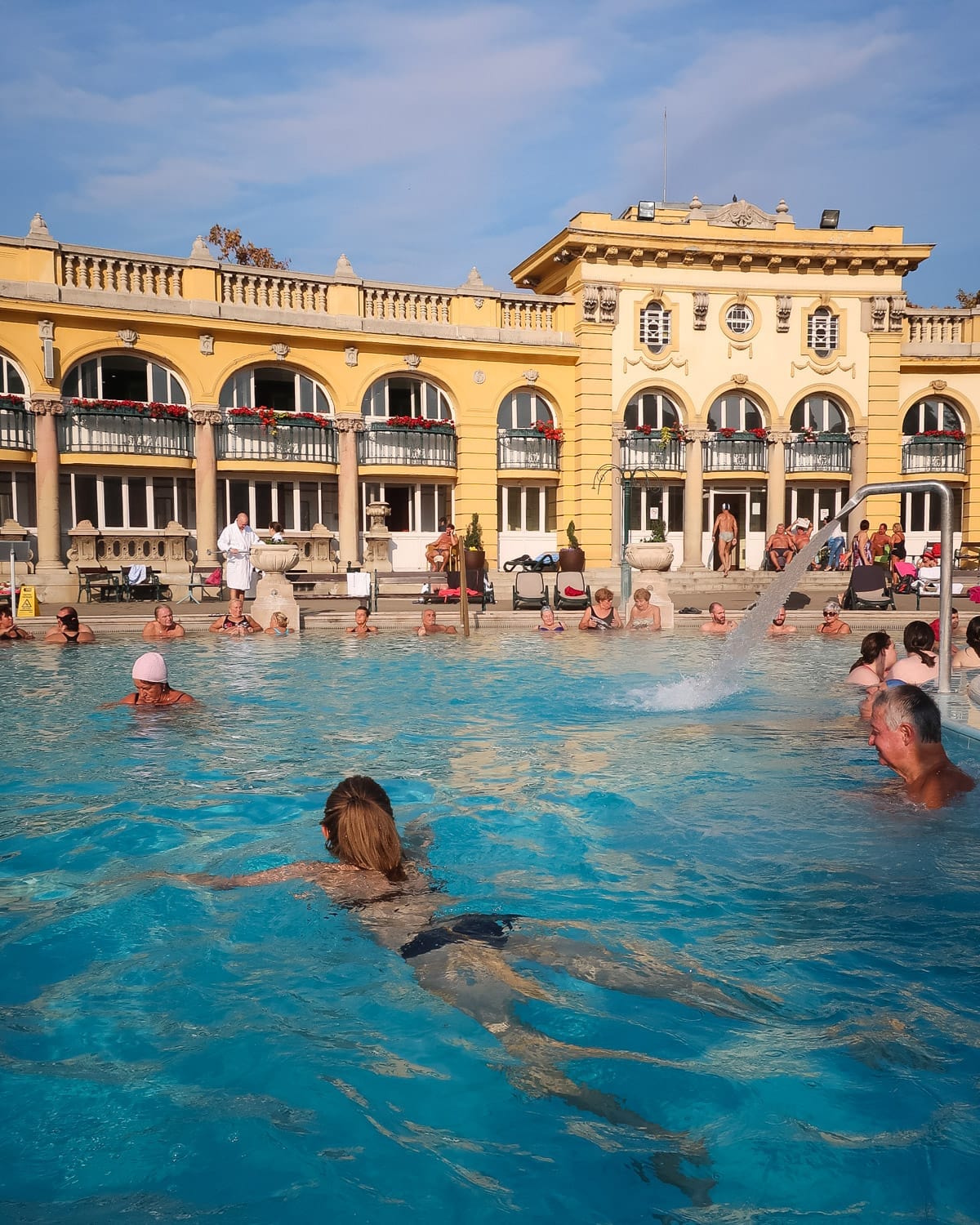 People relaxing in the warm blue waters of the Széchenyi Thermal Bath with yellow neo-baroque buildings in the background.