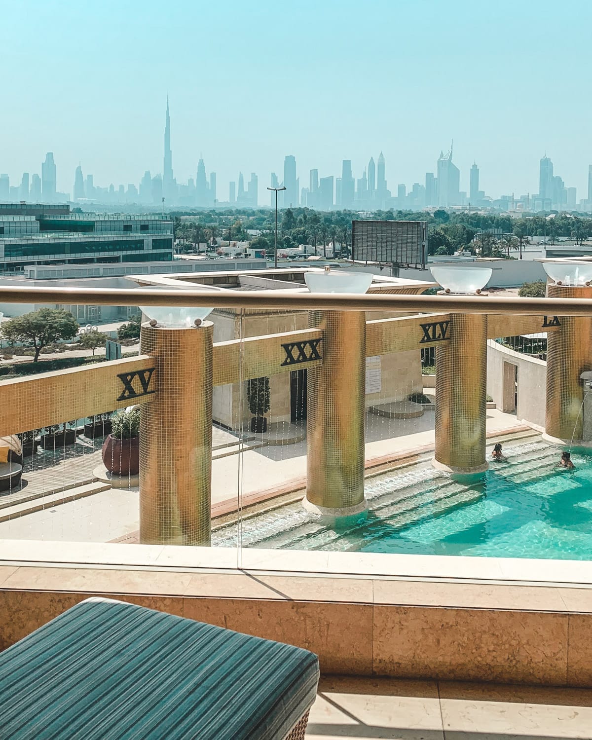 A rooftop pool with golden Roman numerals overlooks the hazy skyline of Dubai, with Burj Khalifa visible in the distance.