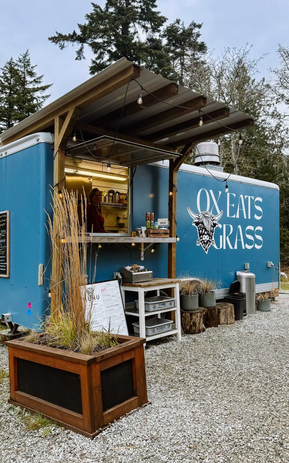 Exterior of the Ox Eats Grass food truck with a blue trailer, string lights, and rustic outdoor setup.