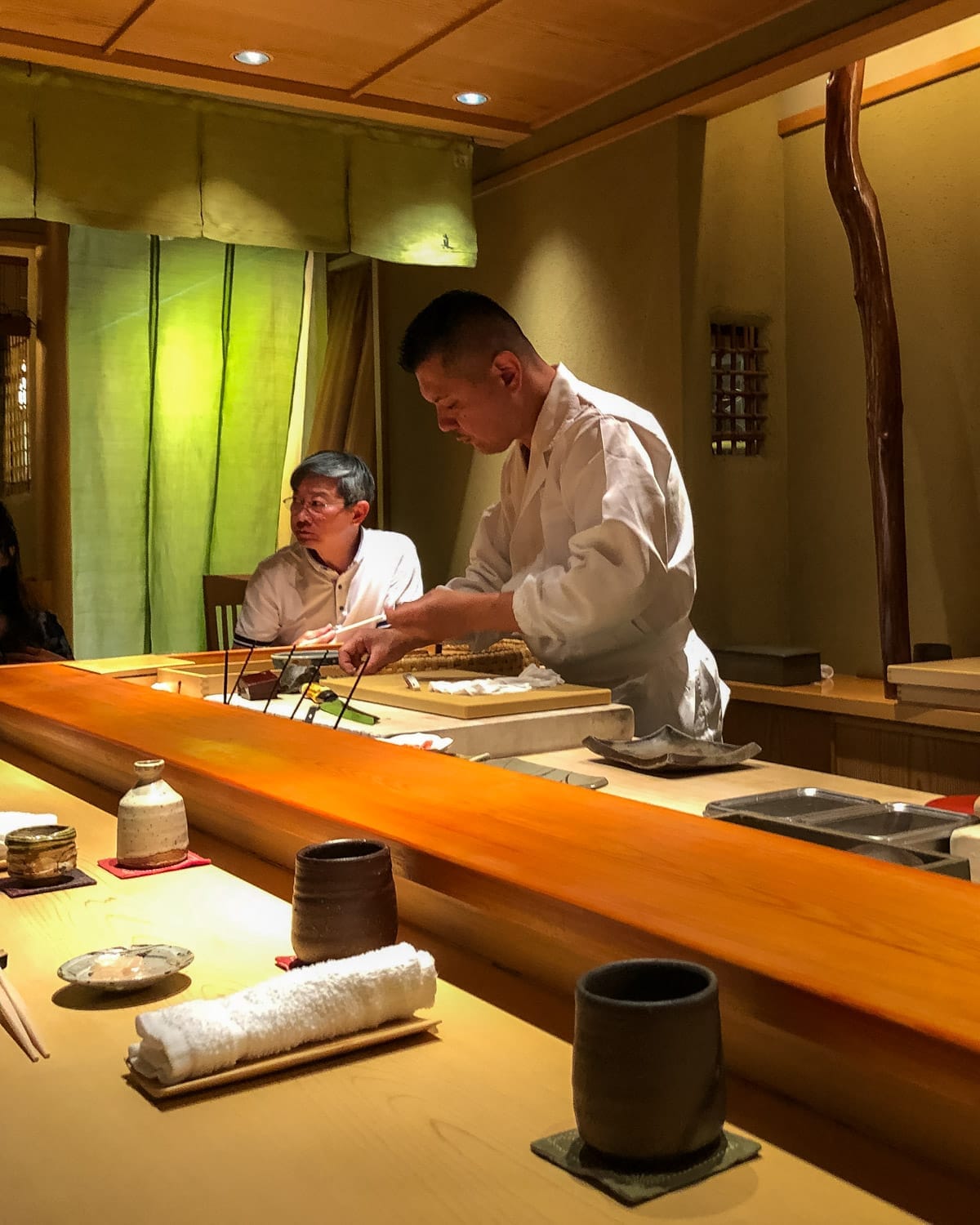 Sushi chef preparing omakase-style dishes behind a wooden counter in an intimate, warmly lit Japanese restaurant.