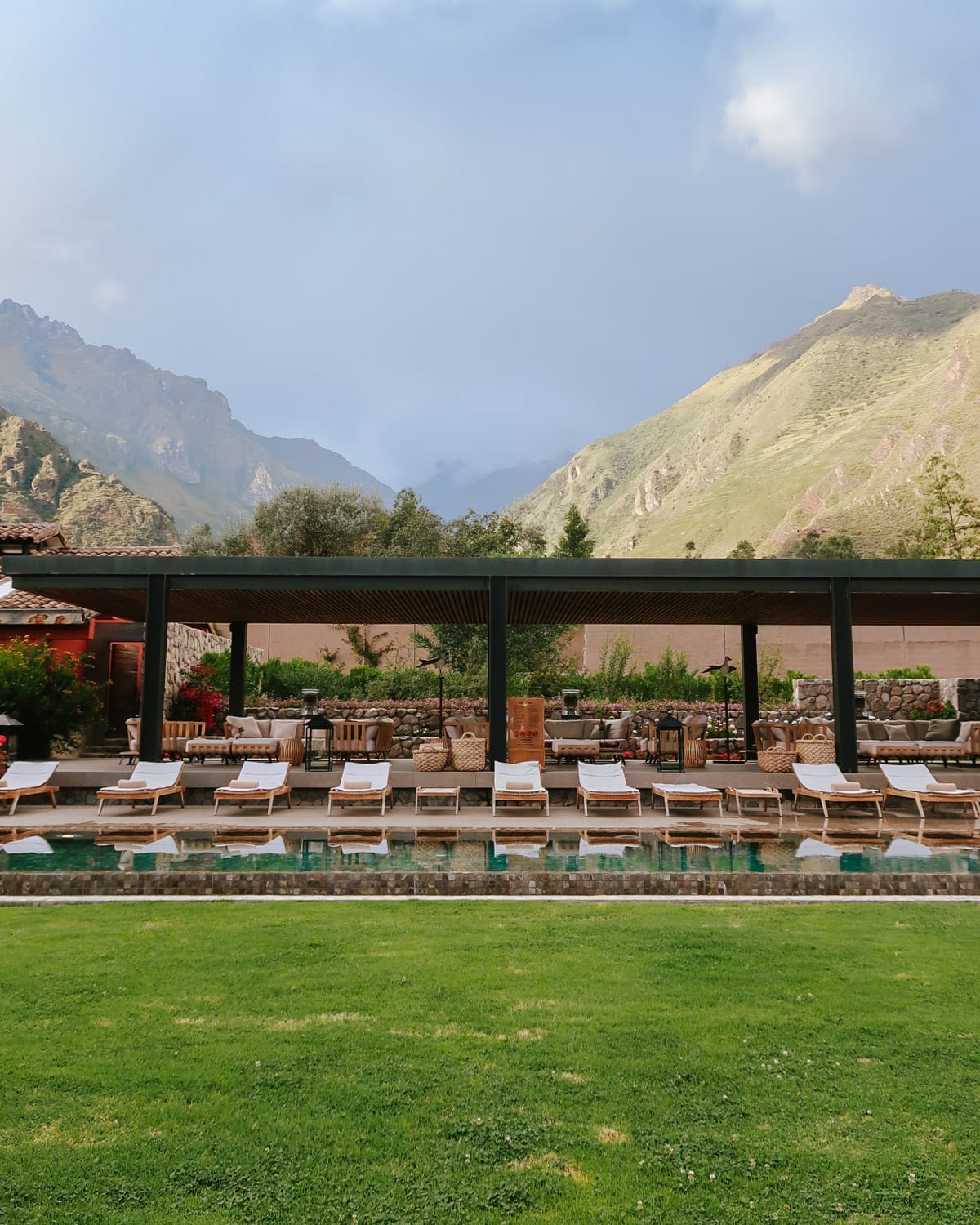 An inviting poolside lounge area with white chairs sits beneath a shaded pergola, backed by green mountains in Peru’s Sacred Valley.