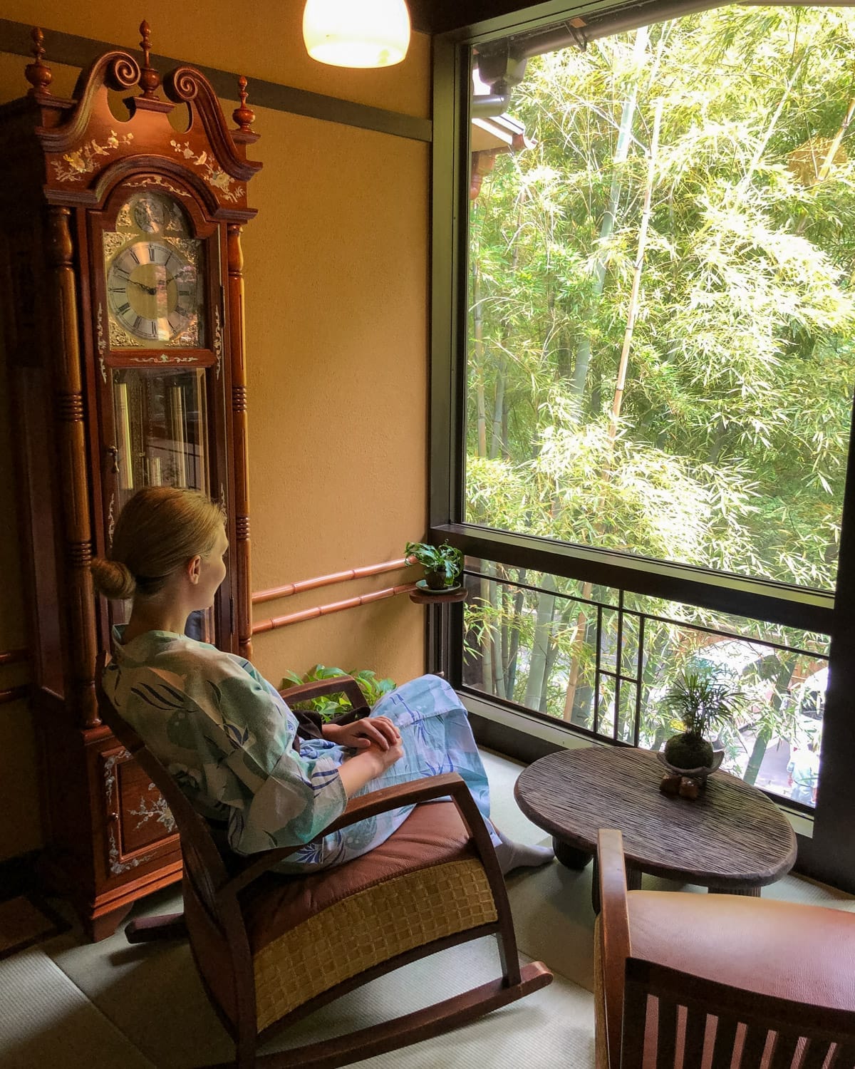 Cec in yukata robe sits in a rocking chair at a traditional Japanese ryokan, gazing out at bamboo through a large window.