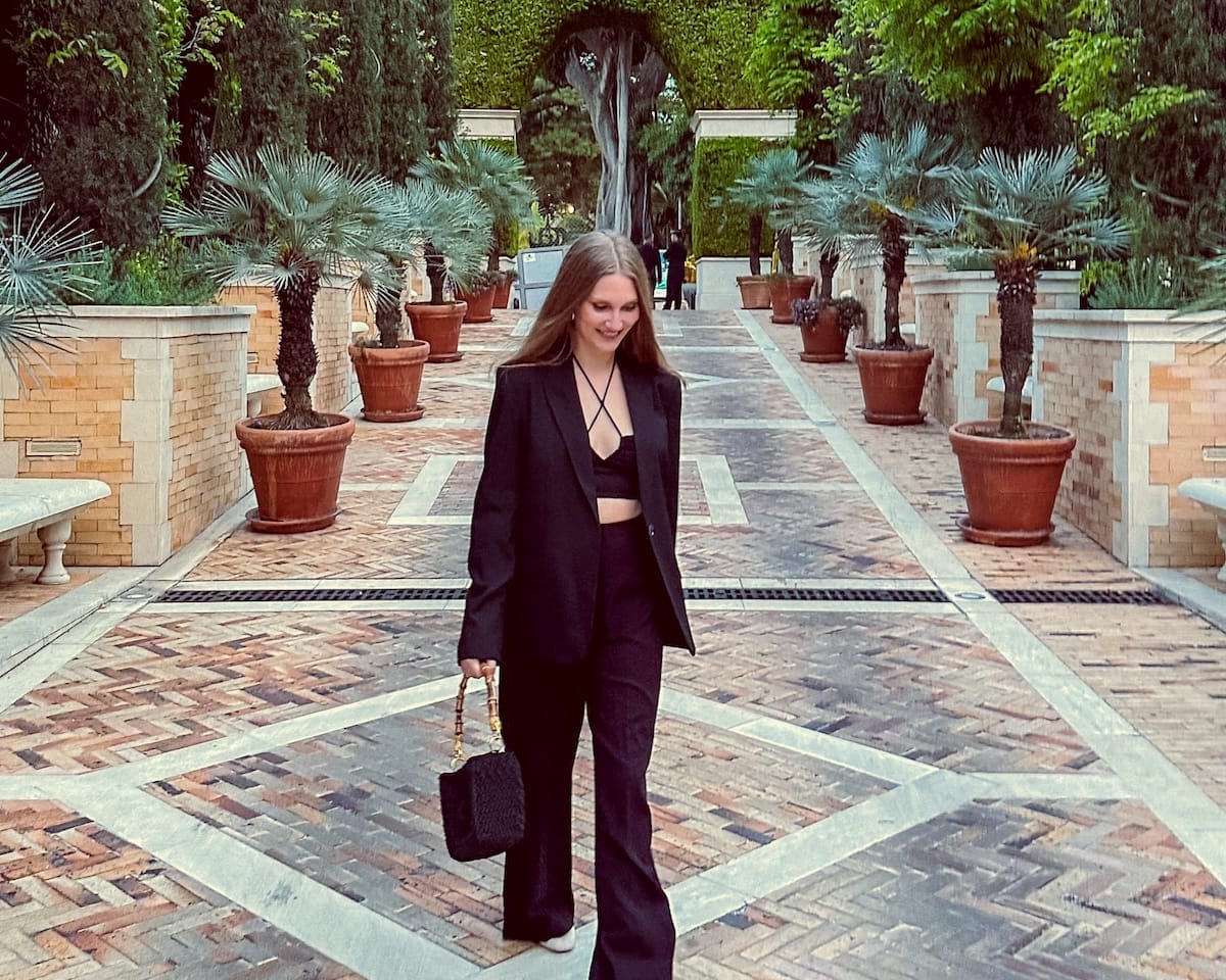 Cec in a stylish black suit walking through a tiled courtyard lined with palm trees and terracotta pots in Monte Carlo.