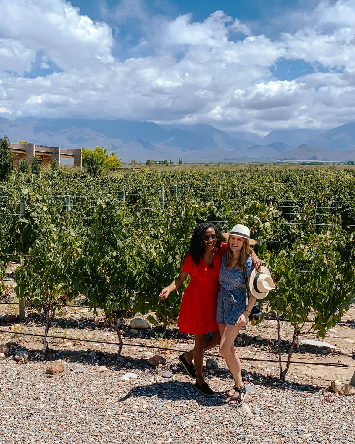 Cec and her friend smiling in a vineyard in Mendoza, Argentina, with the Andes Mountains and blue sky in the background.