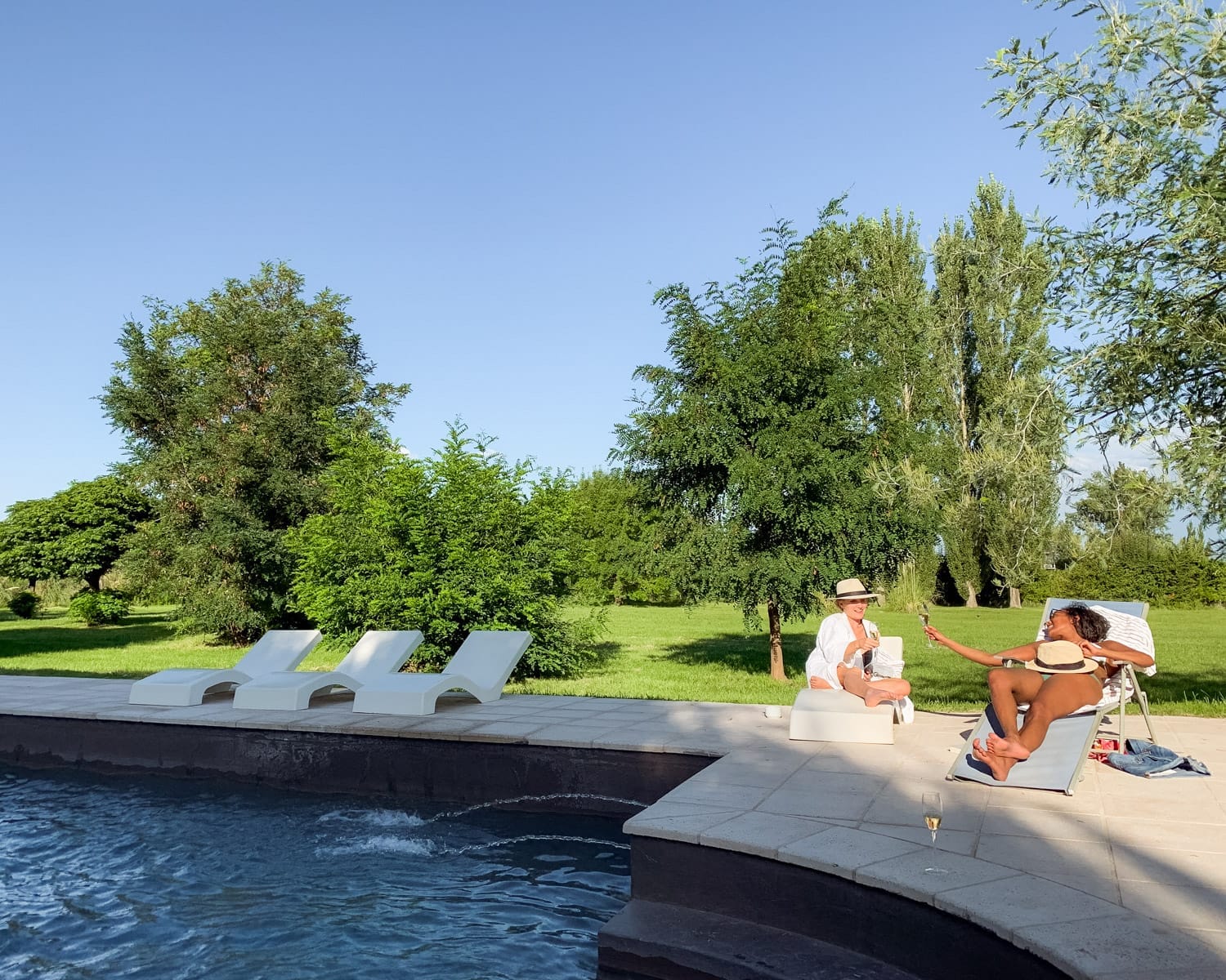 Cec and a friend lounging poolside with drinks on a sunny day at Casa de Huéspedes La Azul, surrounded by green trees and a vineyard estate lawn.