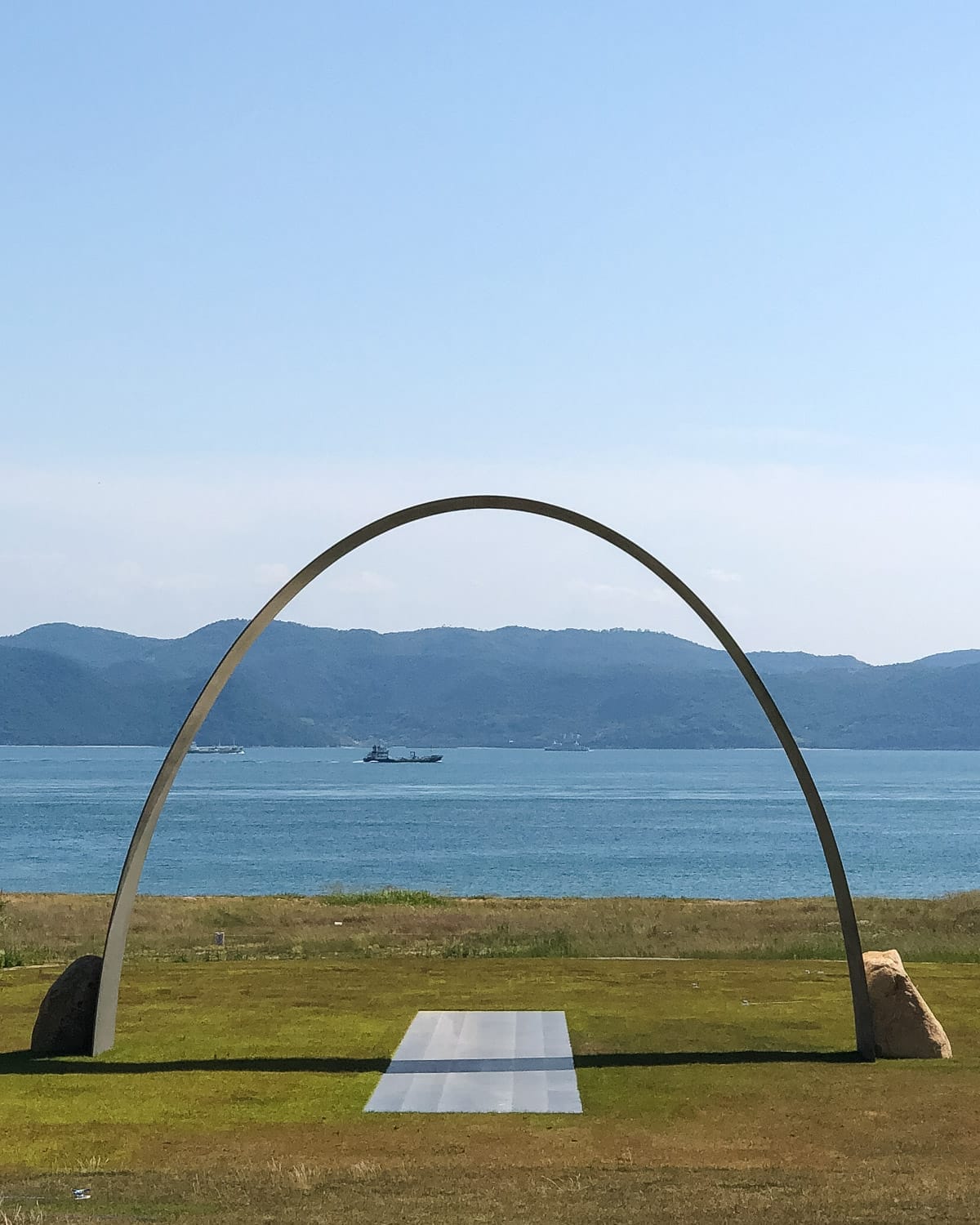 Modern sculpture arch facing the sea on Naoshima Island with calm waters and distant hills.