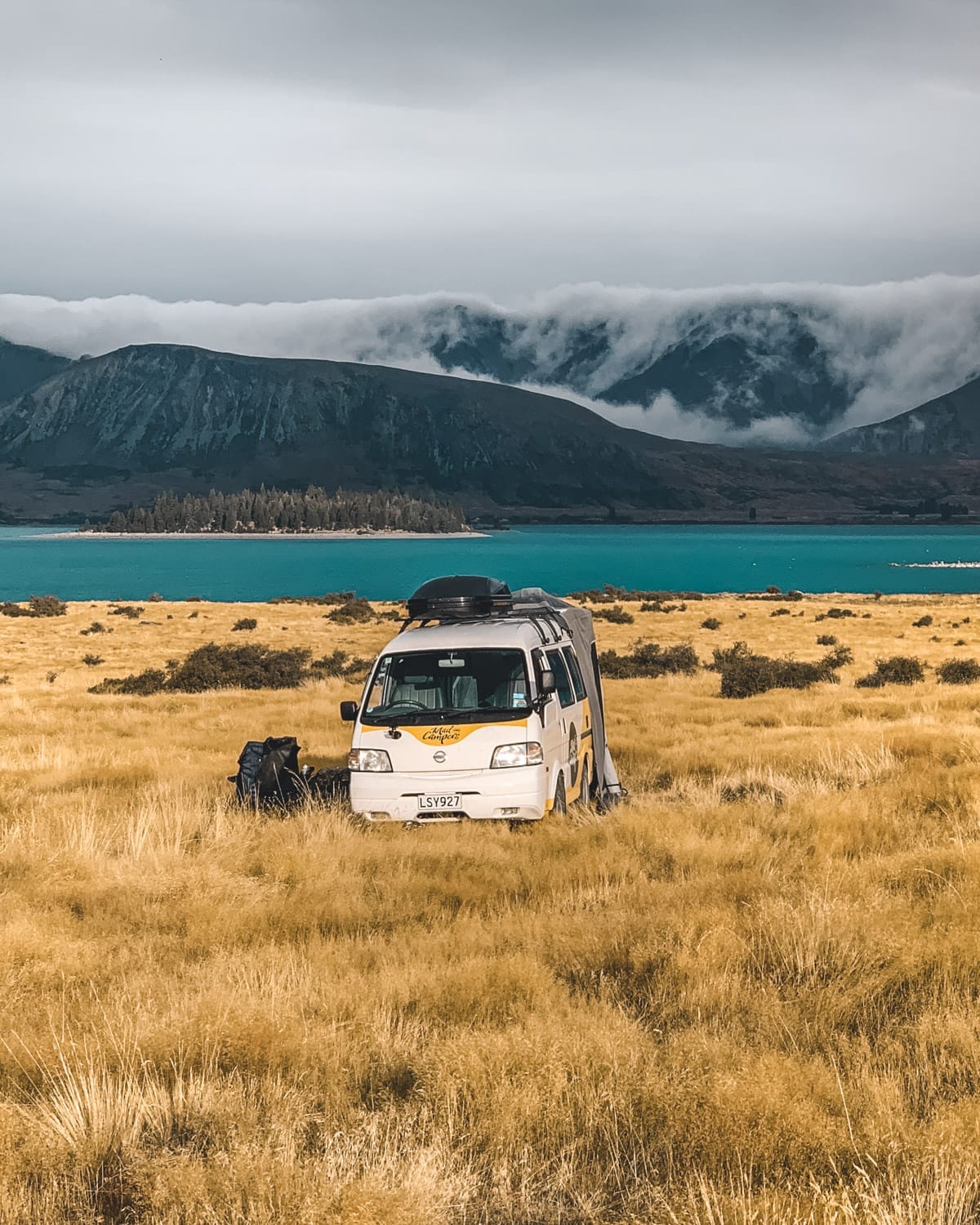 A white and yellow campervan parked in golden grass near a turquoise lake, with misty mountains in the distance on New Zealand's South Island..