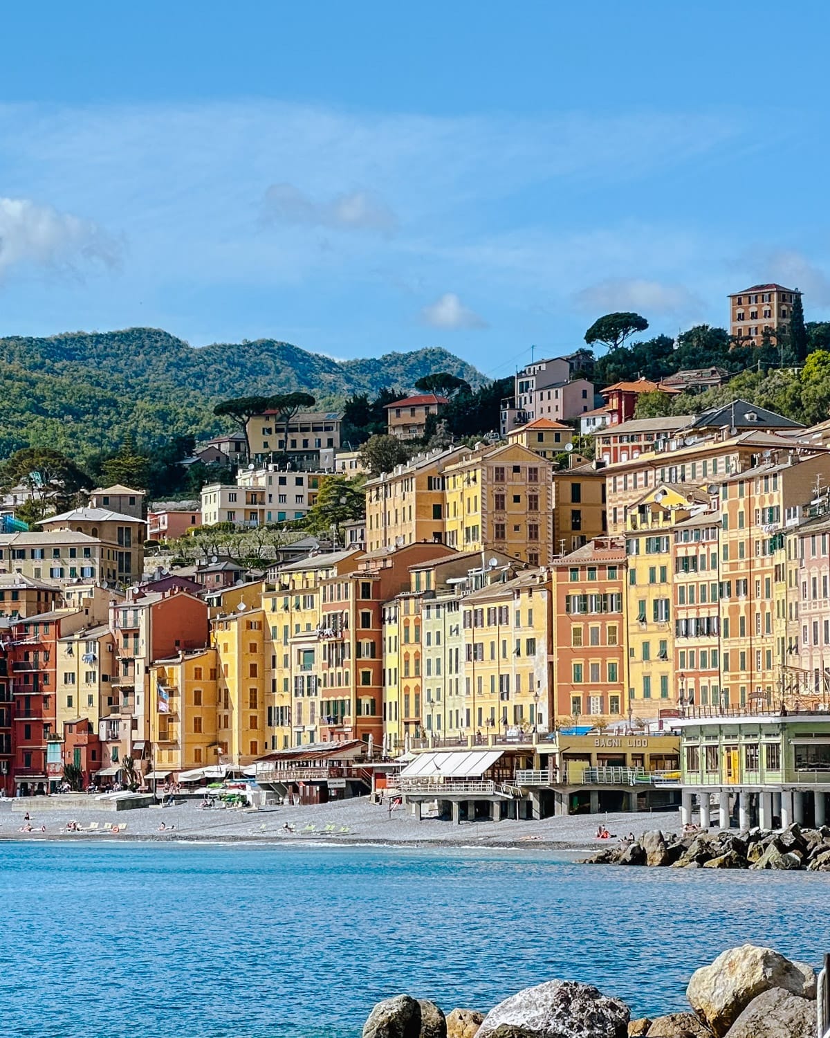 Colourful seaside buildings of Camogli line the beach with green hills rising in the background under a clear sky.