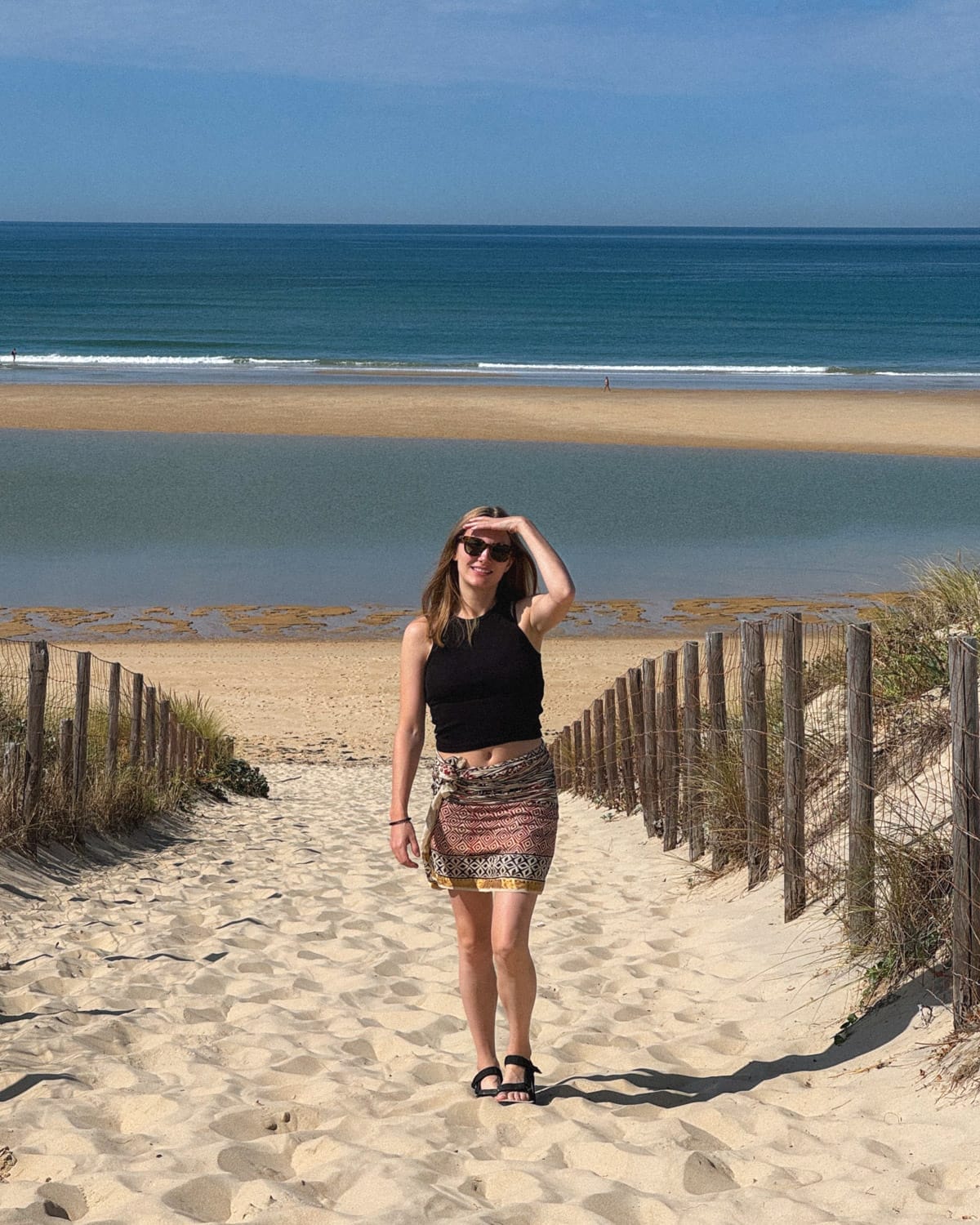 Cec walks along a fenced sandy path away from the vast Atlantic Ocean in Cap Ferret with waves in the distance.